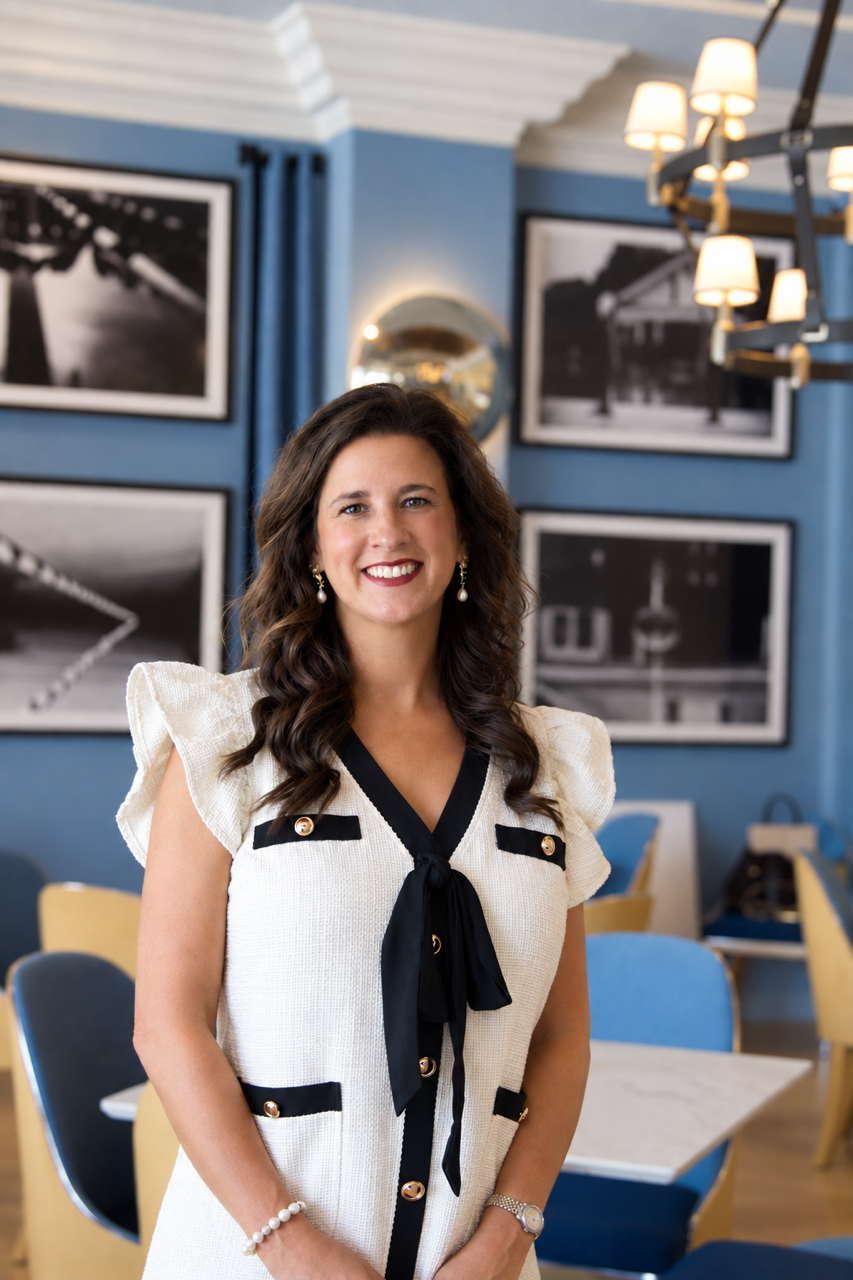 A woman with wavy brown hair smiling in a restaurant or cafe with blue walls and black-and-white photographs, wearing a cream-colored dress with black accents, a black bow tie, pearl earrings, and a watch.