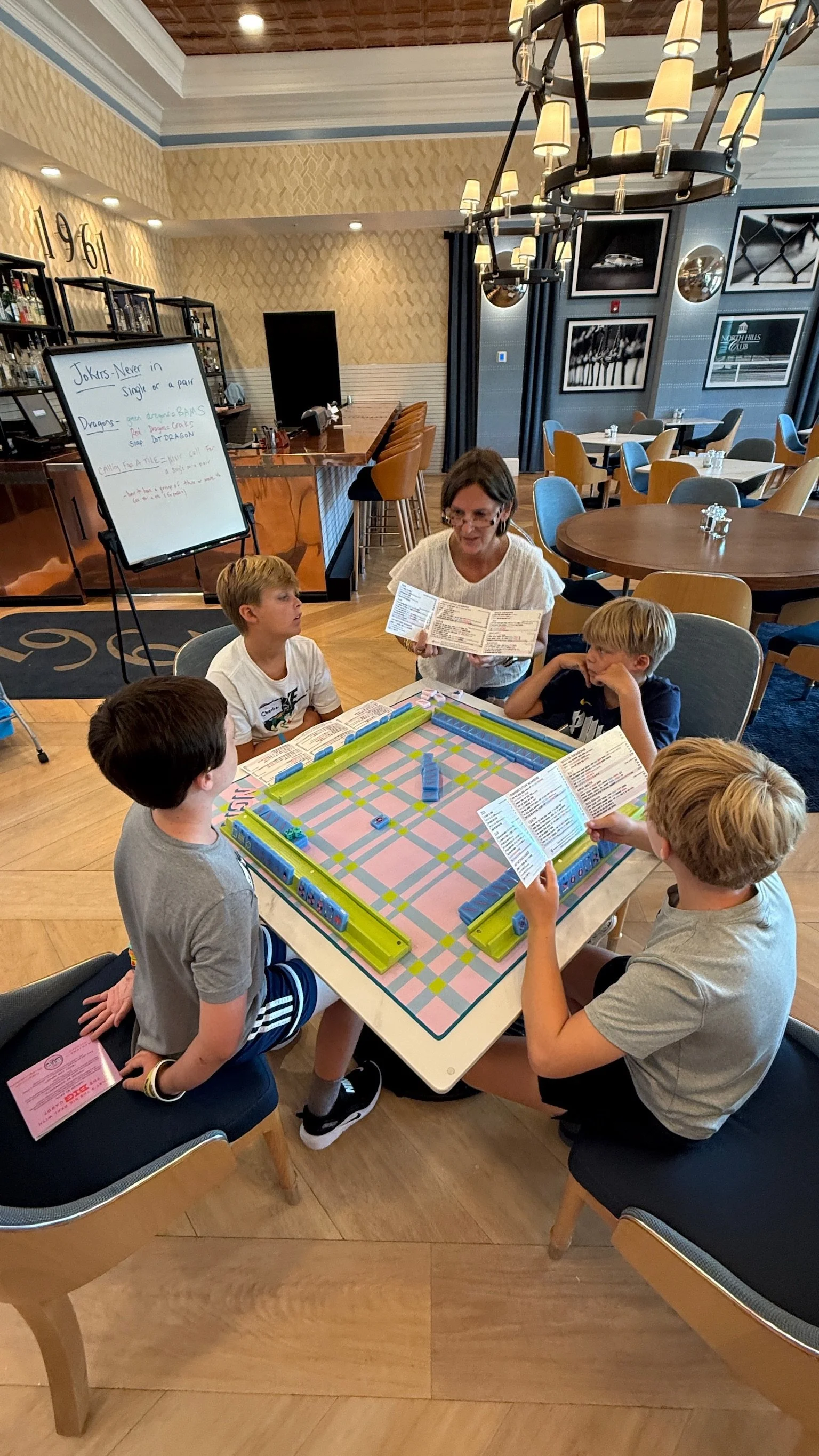 A group of five kids and an adult playing a board game at a table in a restaurant or cafe. The adult woman is holding a menu, and the kids are looking at the game or the woman. The room has a bar area, framed pictures on the walls, and a chandelier hanging from the ceiling.