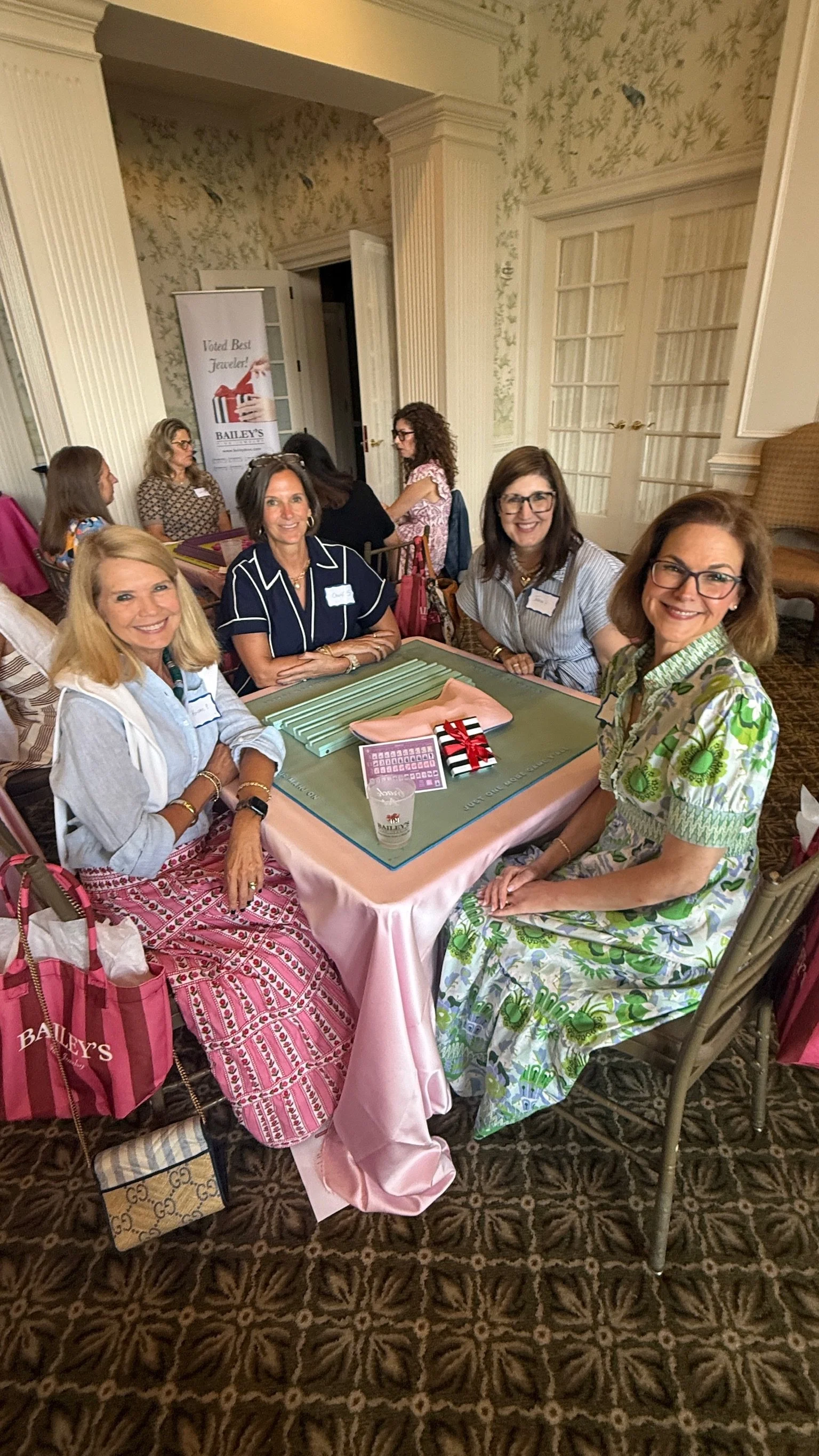 A group of women sitting around a table at an indoor event, smiling at the camera, with gift bags and a pink tablecloth, in a room with cream-colored walls and decorative molding.