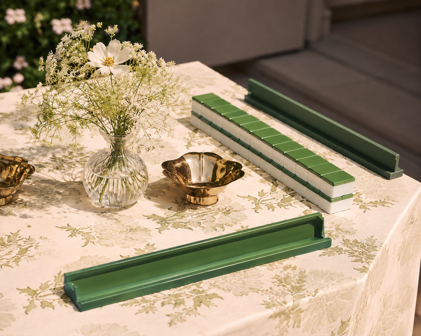 A table with a floral tablecloth, a glass vase with white and cream flowers, and green and white ceramic tiles arranged on the table.