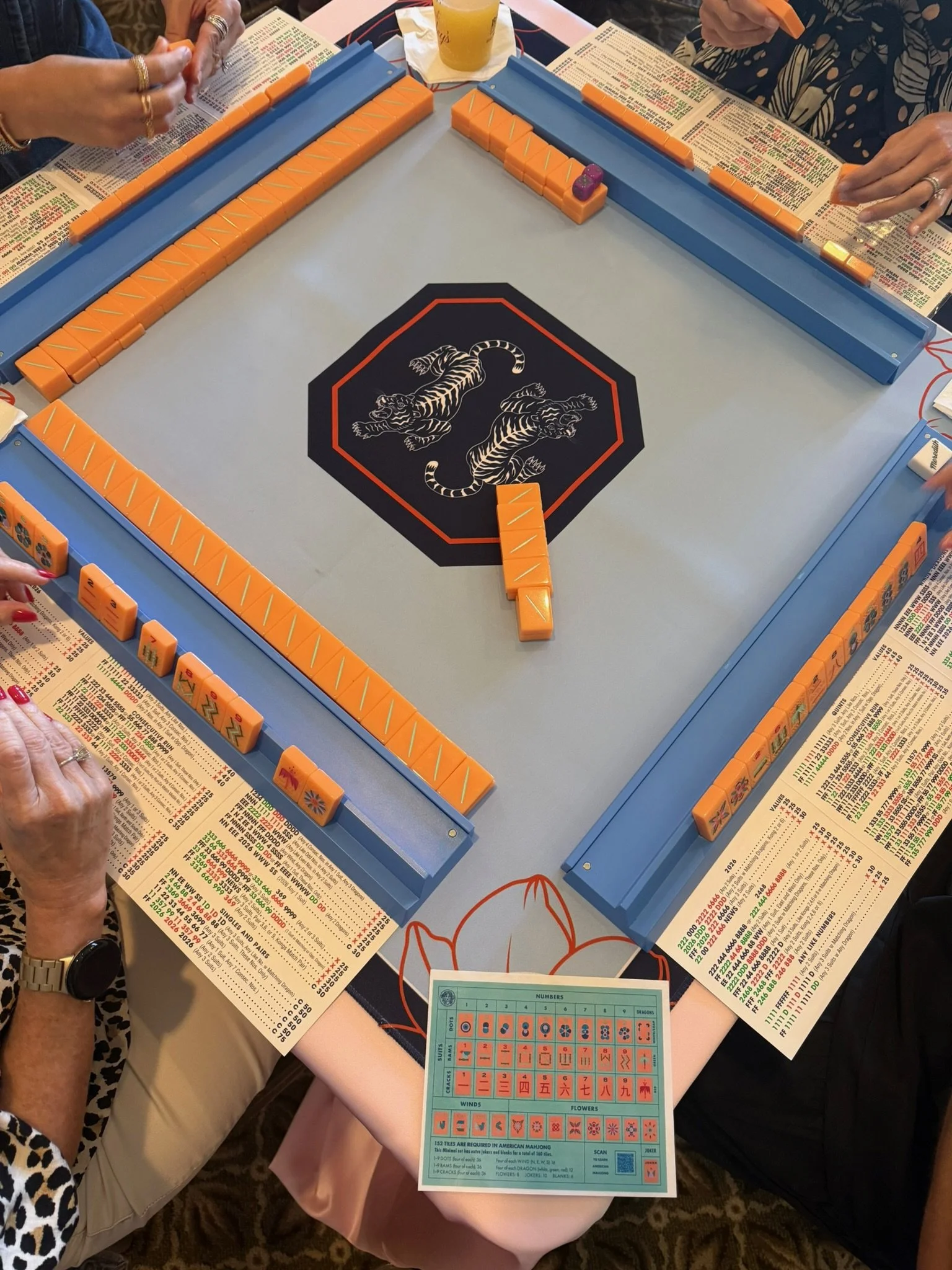 People playing a game of Mahjong at a table with Mahjong tiles, score sheets, and drinks. The table has a central black and red dragon graphic, and a blue scoring sheet is visible.