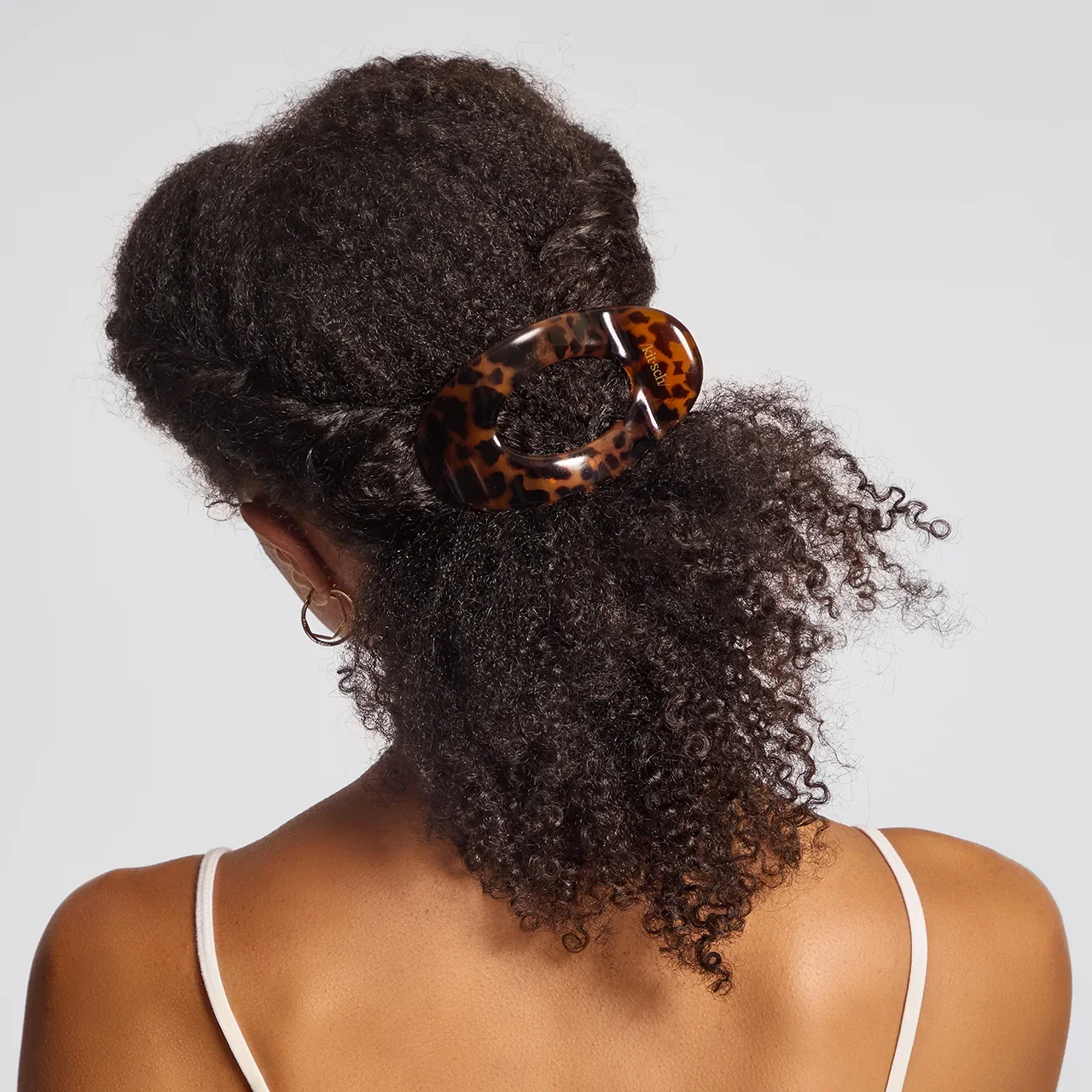 A woman with curly dark hair in a half-up, half-down hairstyle secured with a large tortoise shell hair clip, model's bare shoulder and white strap top visible, neutral background.