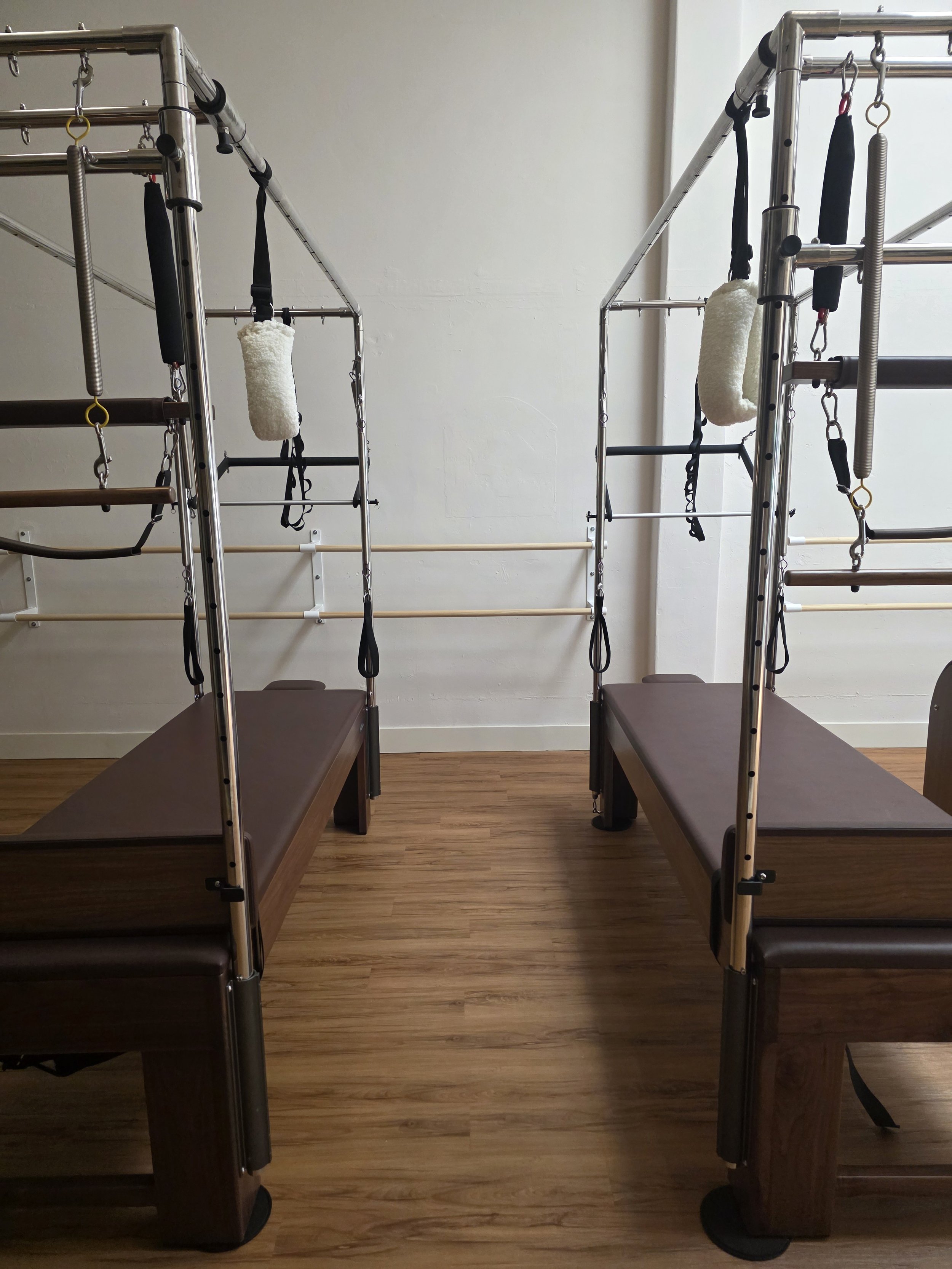 Pilates Cadillacs with walnut platforms and chrome metal frames in a fitness studio with white walls and wood-style flooring.