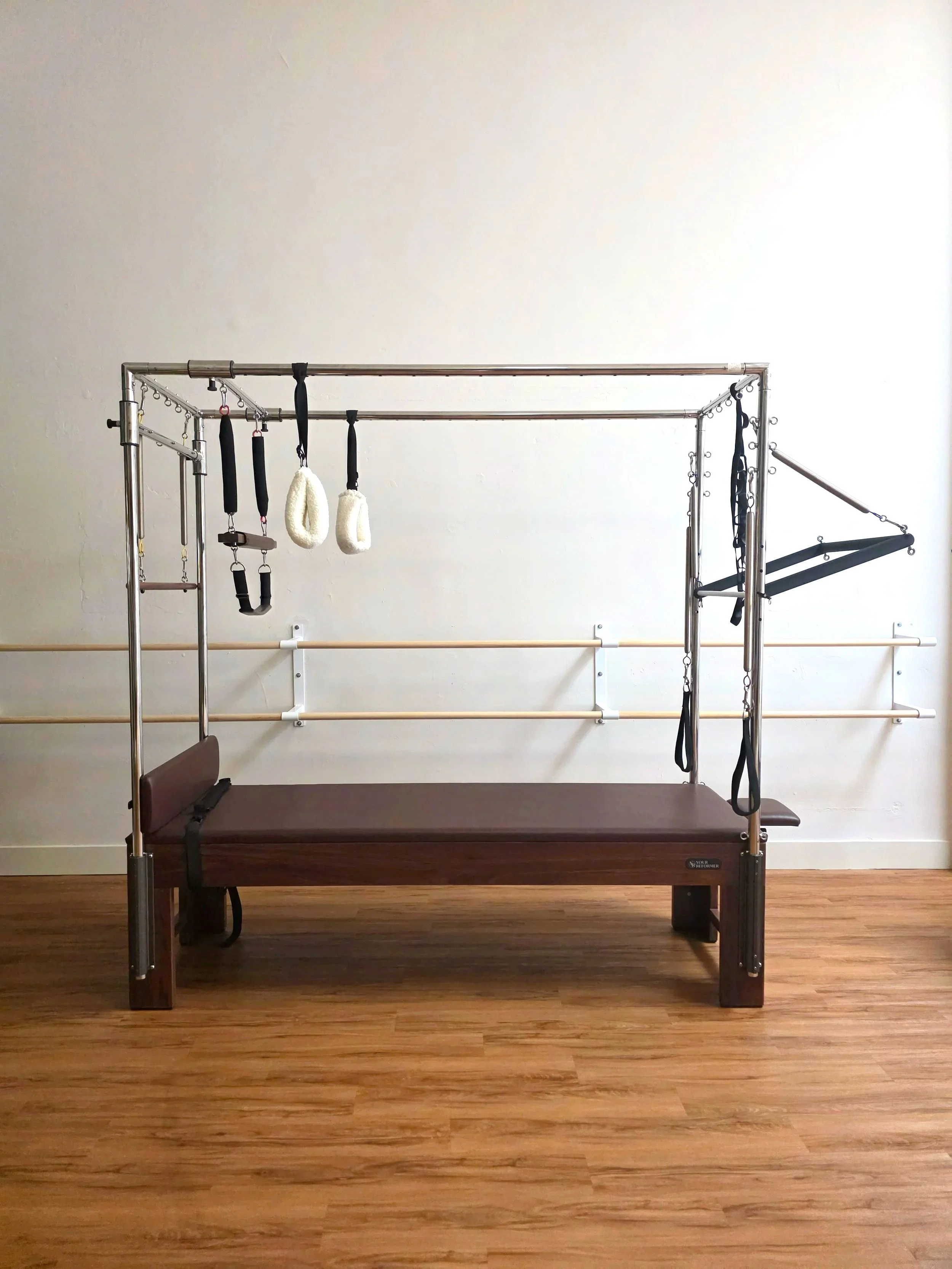 A Pilates Cadillac with various straps, handles, and a cushioned platform, set against a plain white wall with ballet barres and hardwood flooring.