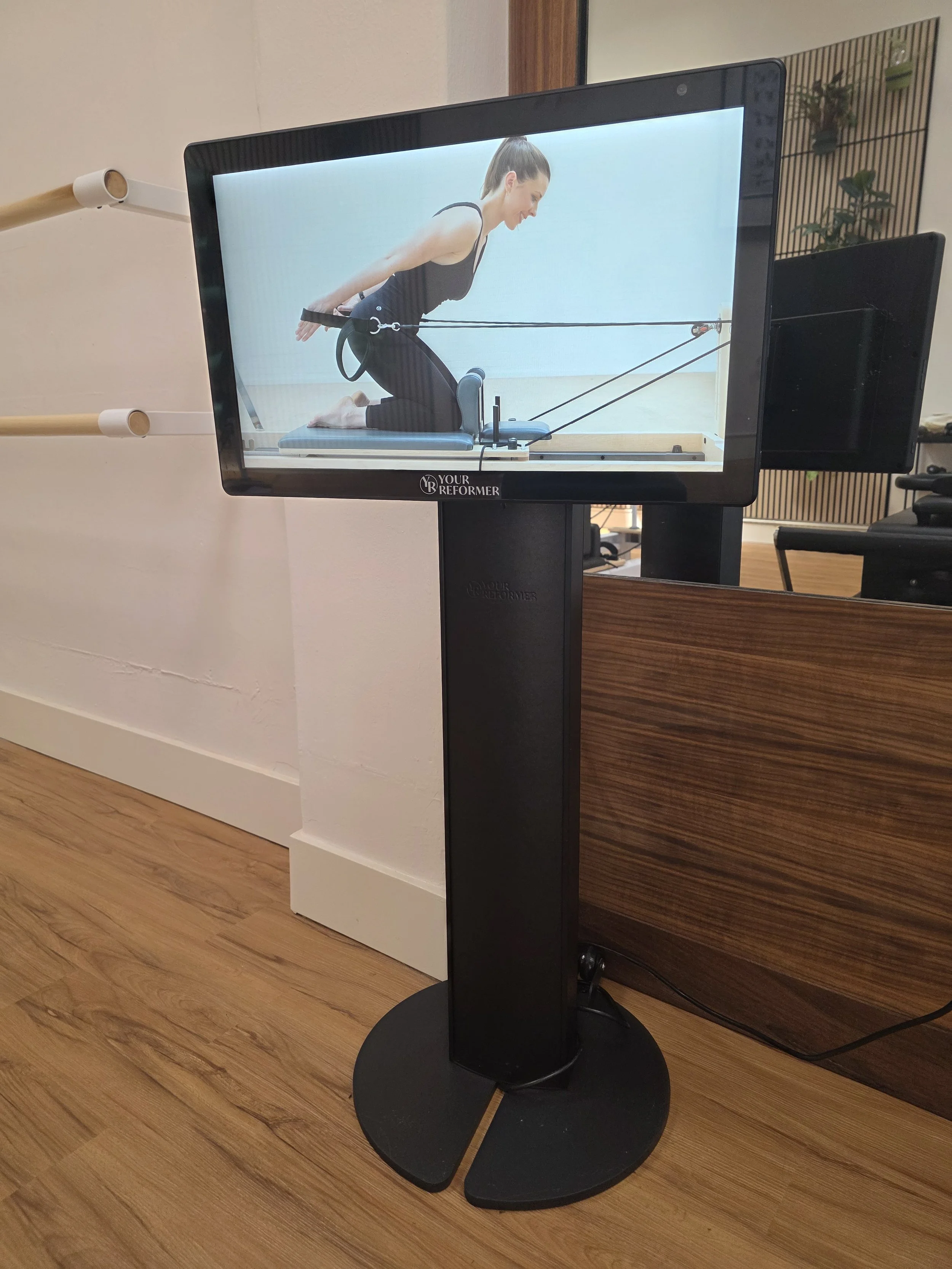 Kiosk: digital screen on a 3 foot tall base featuring a picture of a woman in black workout clothes on a reformer Pilates machine, performing a core and arm exercise on a padded platform with resistance straps. indoors near a mirror and wooden floor.