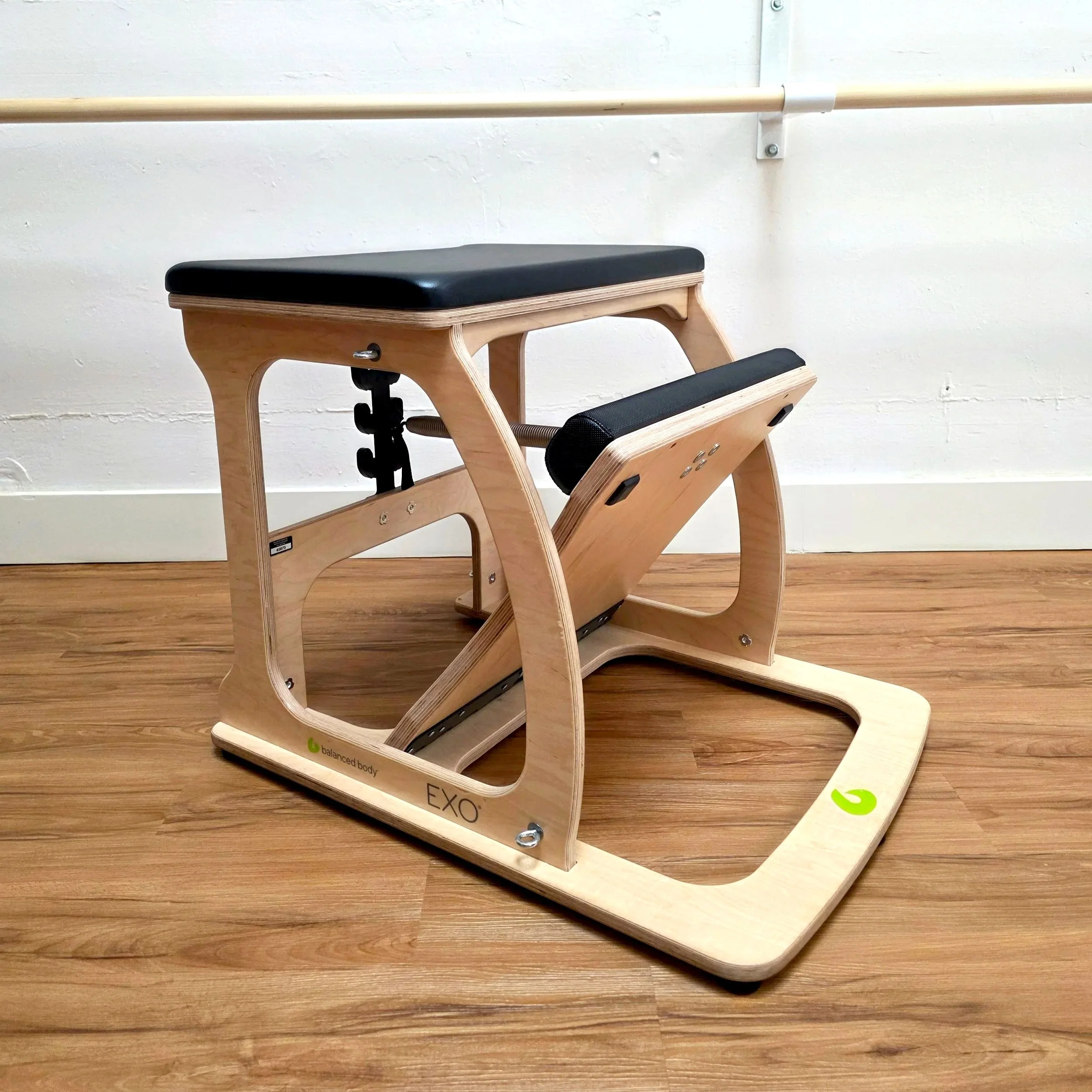 Oak Pilates chair with black padding on a wooden floor against a white wall with ballet barres on it.