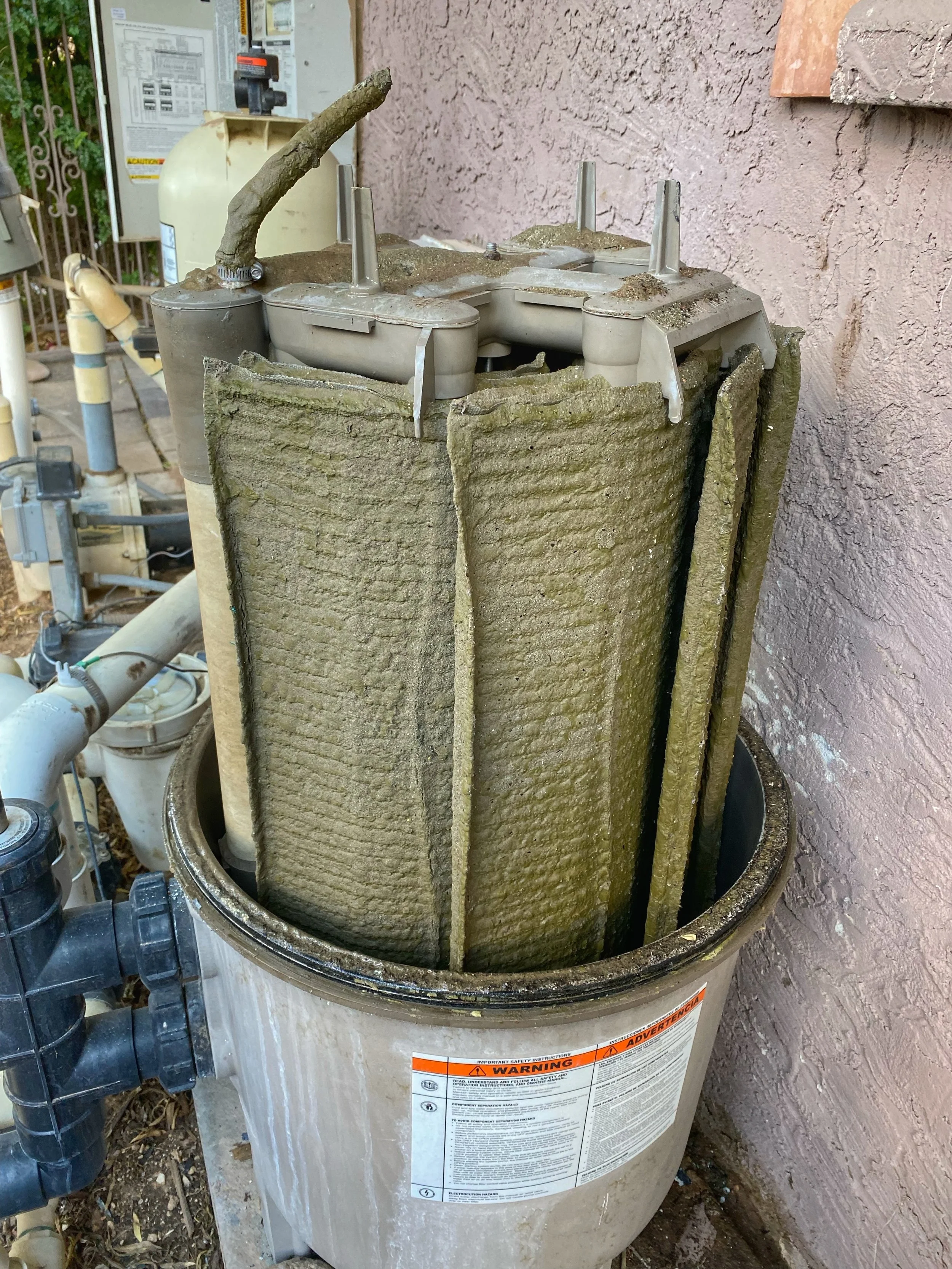 A water heater with the insulation layer removed, exposing the inner heating element and piping, situated outdoors next to a textured wall and electrical breaker box.
