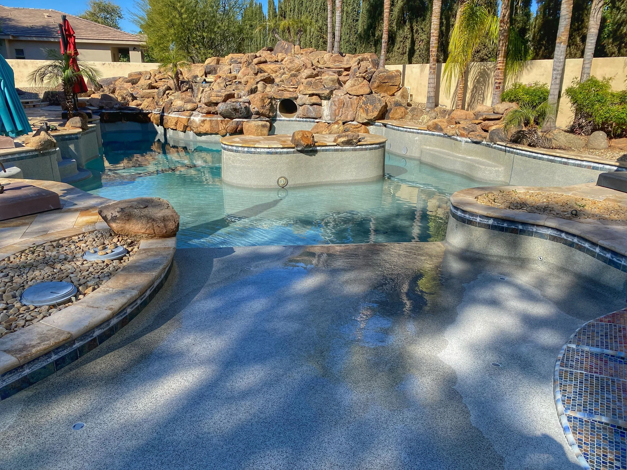 A backyard swimming pool with a waterfall feature surrounded by rocks and lush greenery, including palm trees and bushes, with a house in the background.