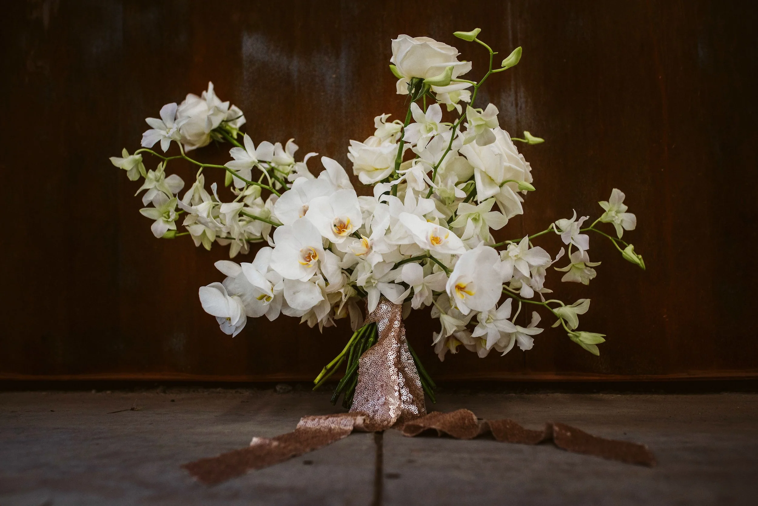 An organic and sculptural wedding bouquet made of pristine white Phalaenopsis orchids, Dendrobium orchids, and roses, tied with a shimmering rose gold sequin ribbon, resting on a concrete floor against a rustic oxidized metal background.