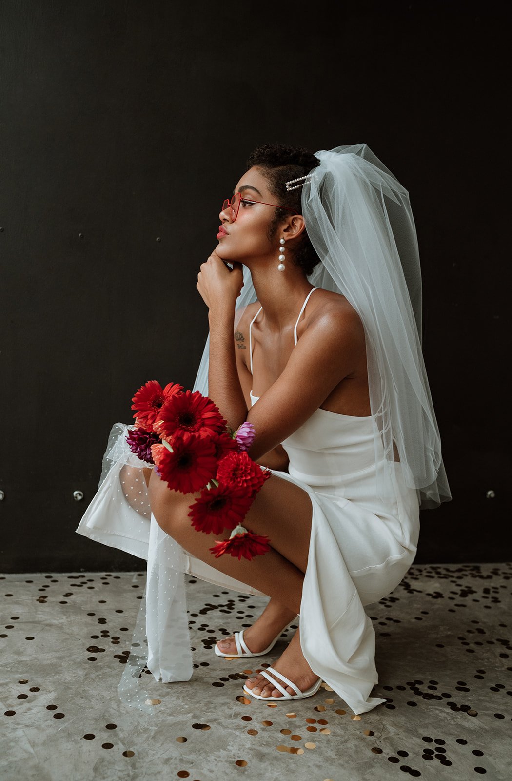 Profile portrait of a bride crouching while holding a vibrant red gerbera daisy bouquet. She wears a minimalist white wedding dress,a tulle veil, large pearl drop earrings, and red-rimmed sunglasses,