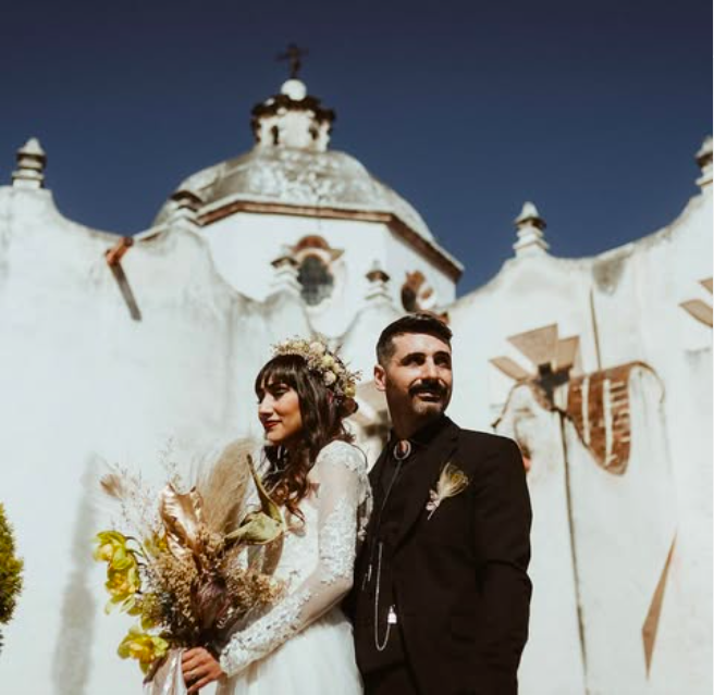 Dramatic low-angle portrait of an alternative wedding couple holding a dried floral bouquet in front of an ancient stone church dome.