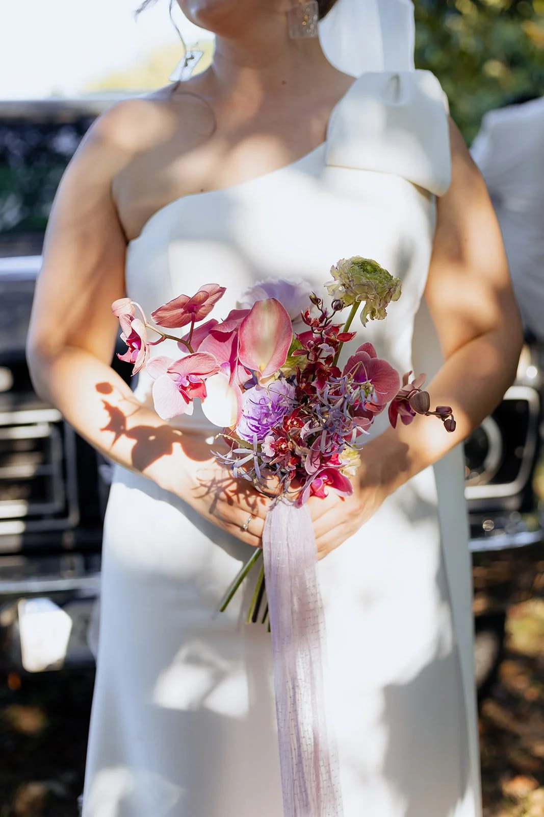Midsection of a bride holding a modern pink and purple floral bouquet with a classic vintage car in the background.