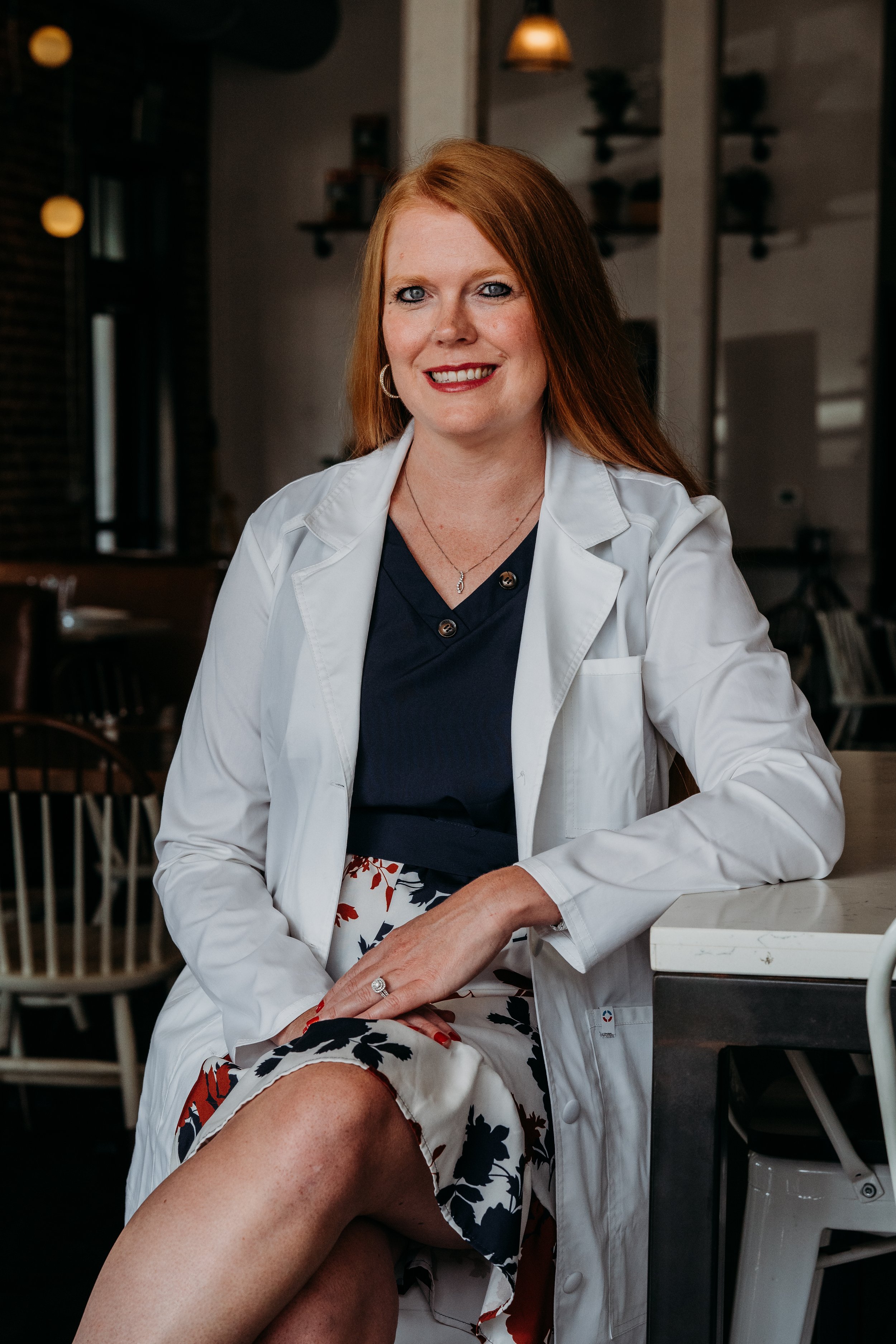 A woman with red hair and blue eyes smiling, wearing a white coat and navy top, sitting at a table in a cafe or restaurant.