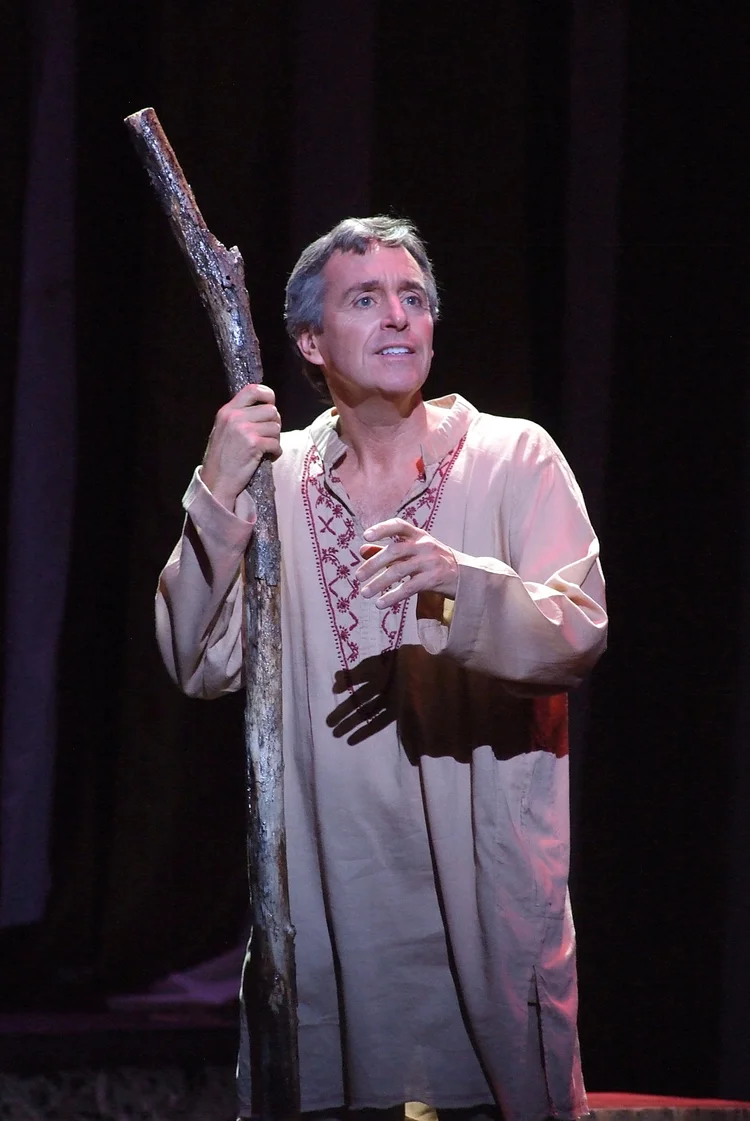 A man in a beige garment holding a large stick on a dark stage.