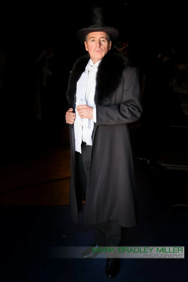 Person dressed in classic theatrical attire, including a tall top hat, black fur collar coat, white scarf, and black shoes, standing in a dark setting.