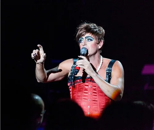 Person dressed in drag, with makeup and a pearl necklace, singing into a microphone on stage.