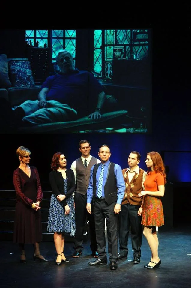 Stage scene with six actors standing in a row on stage. A large projected image of an older man sitting on a couch with a window behind him is in the background.