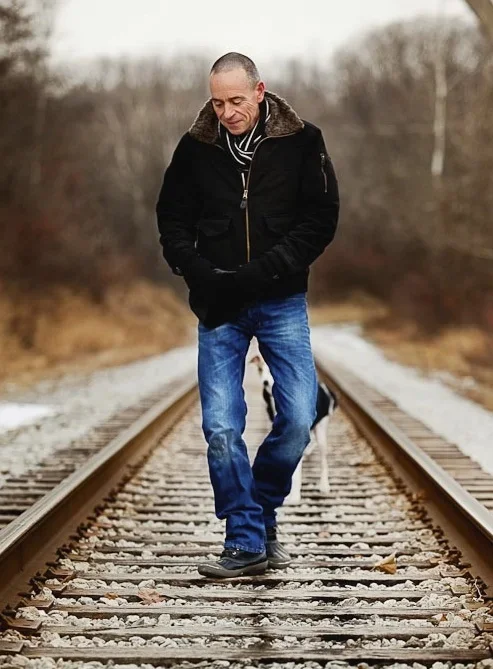 Man walking on train tracks outdoors during autumn or winter, wearing a black jacket, blue jeans, and black shoes.