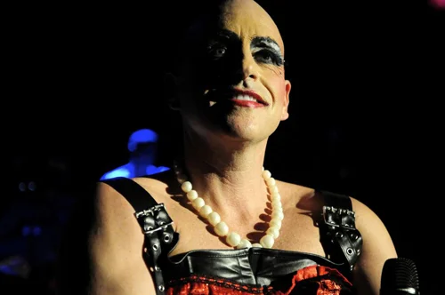 Person dressed in drag with makeup, pearl necklace, and a costume, smiling at the camera.