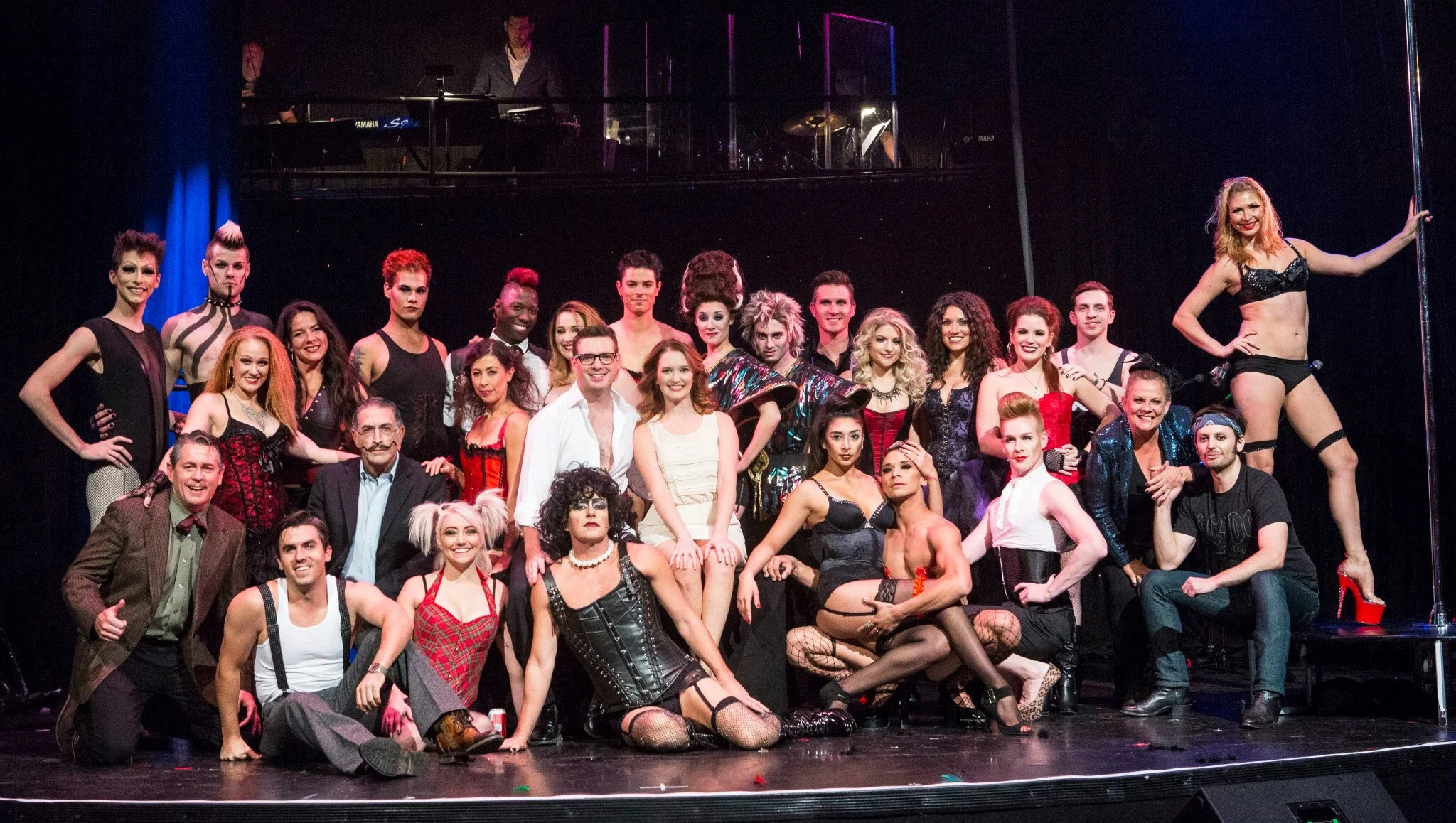 Group of people on stage, some in costumes, posing for a photo after a performance, with a pianist in the background on an upper level.