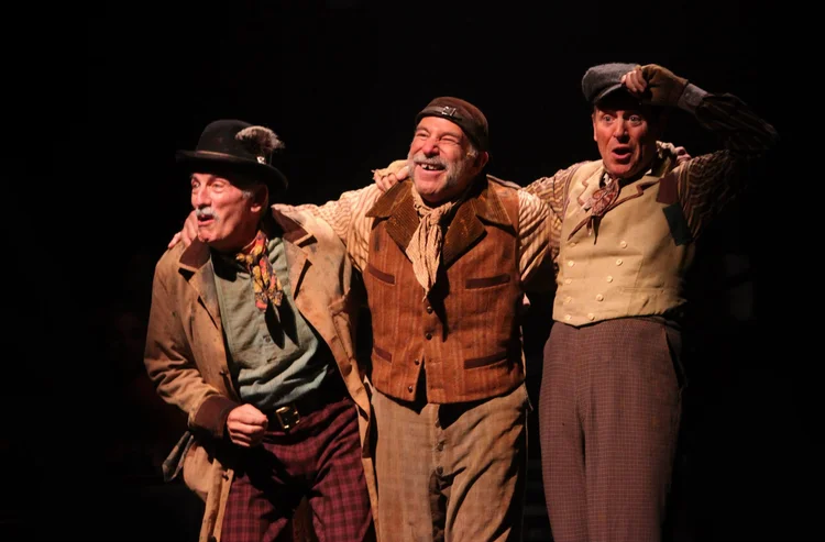 Three actors in vintage costumes are happily taking a bow on stage, with arms around each other.