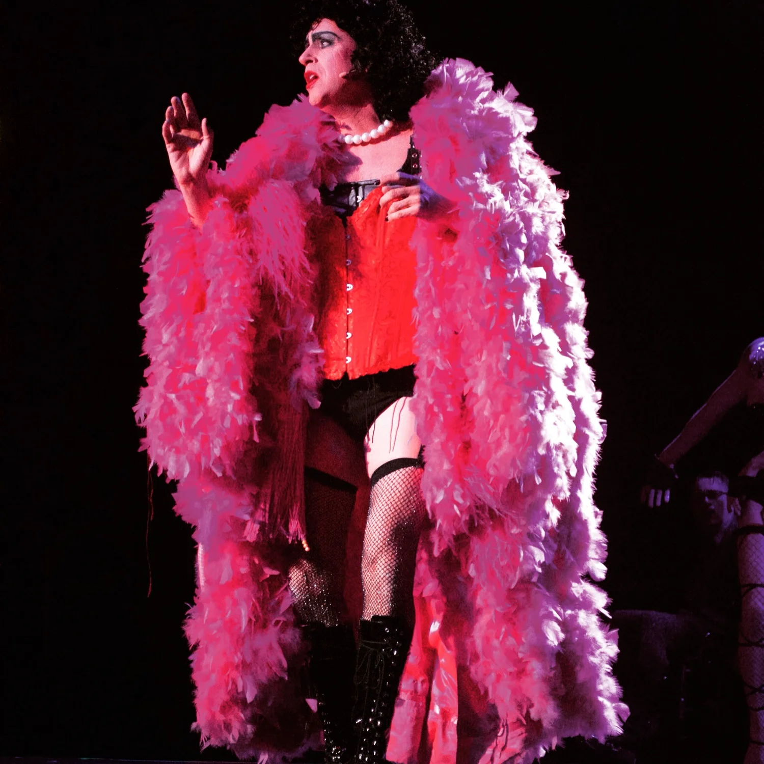 Performer dressed in a red corset, fishnet stockings, thigh-high boots, and a fluffy pink feather boa, holding a microphone, on stage with dark background.