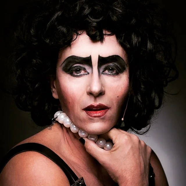 Close-up portrait of a person with dramatic eye makeup, curly black hair, red lipstick, wearing a pearl necklace and a black strap top, with hand resting on chin.