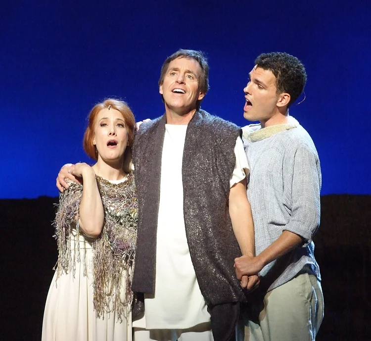 Three actors performing on stage, with a dark background. The woman has red hair and wears a patterned shawl, singing with a worried expression. The man in the center has gray hair and a vest, singing passionately. The young man on the right, with sh