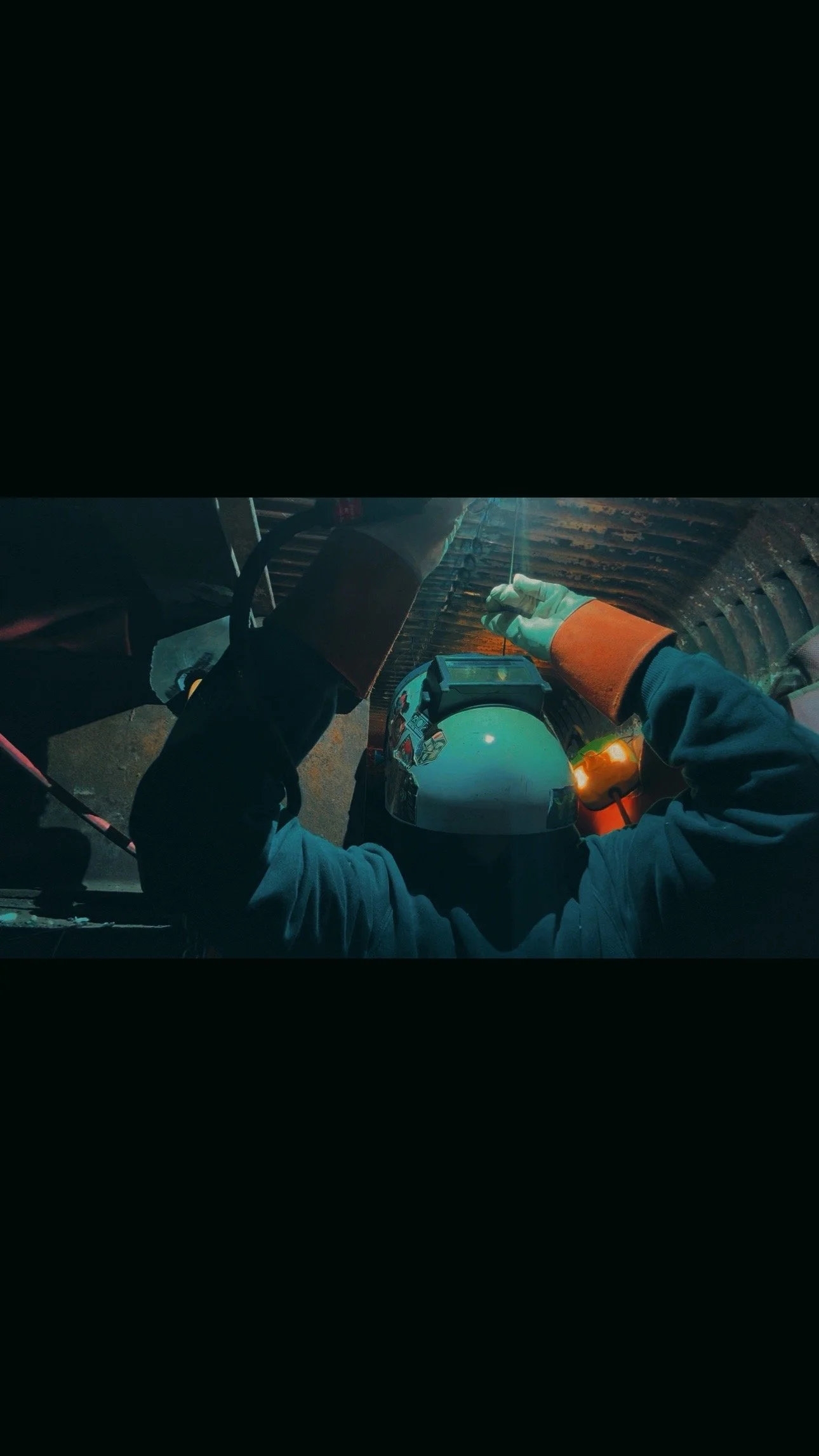 A person welding with a welding helmet and gloves inside a tunnel or confined space, working on metal structure.