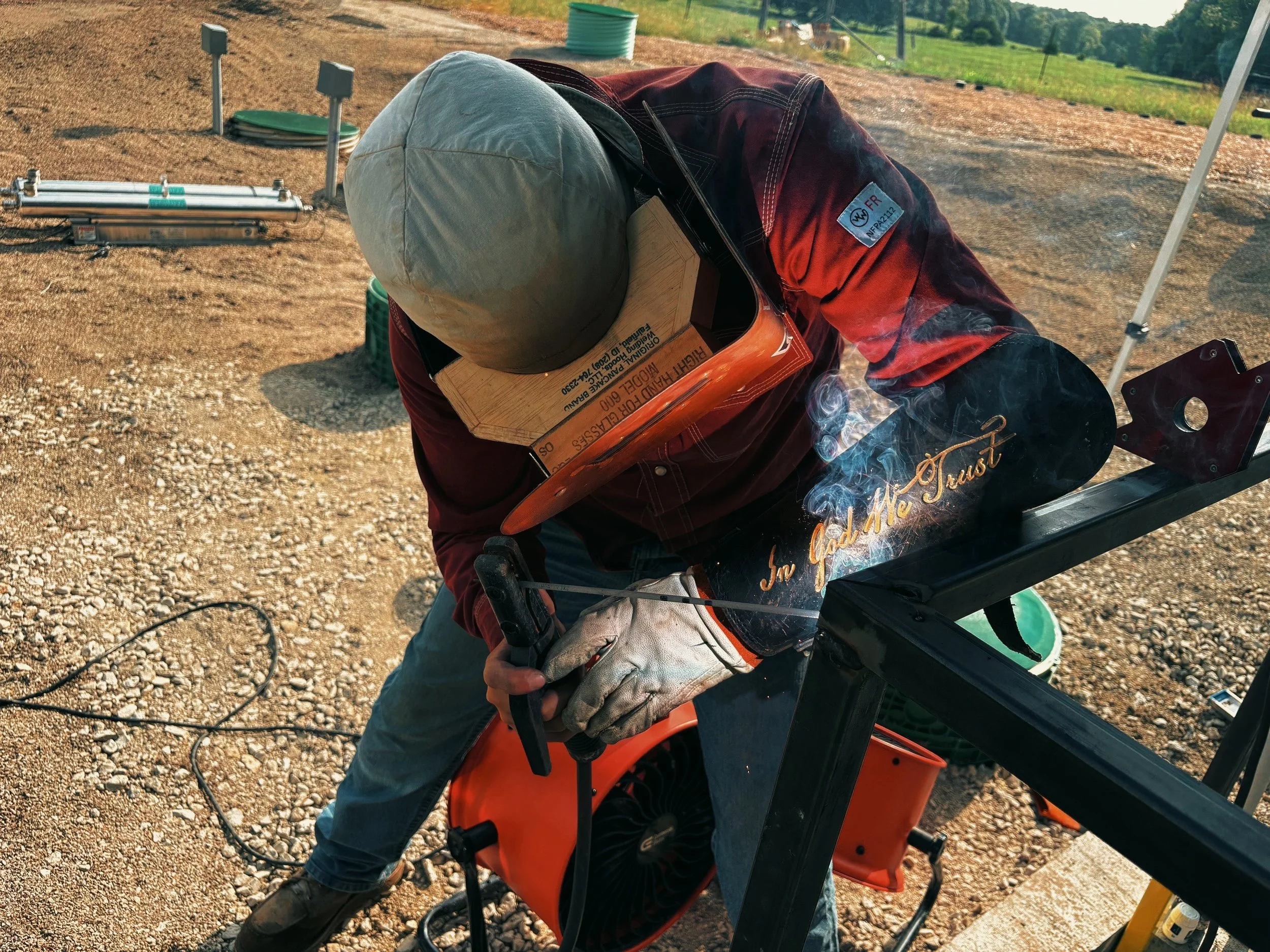 A person wearing a protective helmet, gloves, and a red plaid shirt is working outdoors on heavy machinery, possibly welding or performing maintenance. The background shows open land and some farming equipment.
