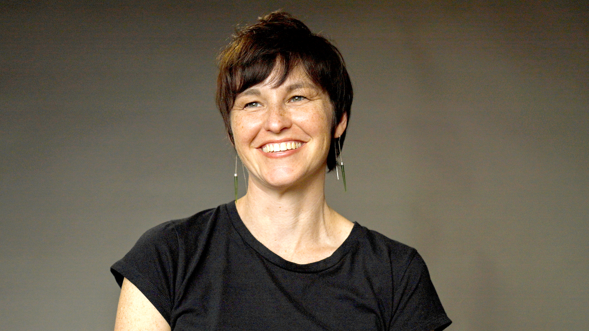 A woman with short dark hair, smiling, wearing earrings and a black shirt against a gray background.