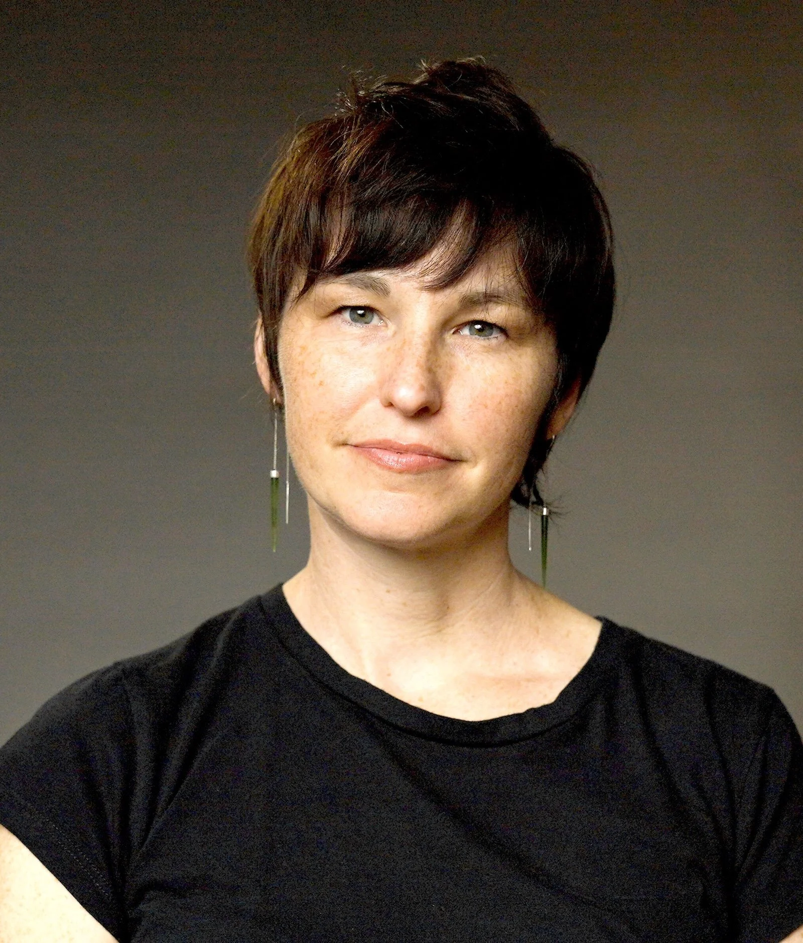 A woman with short dark hair with reddish highlights, light skin, and blue eyes, wearing a black shirt and earrings, smiling slightly against a neutral background.