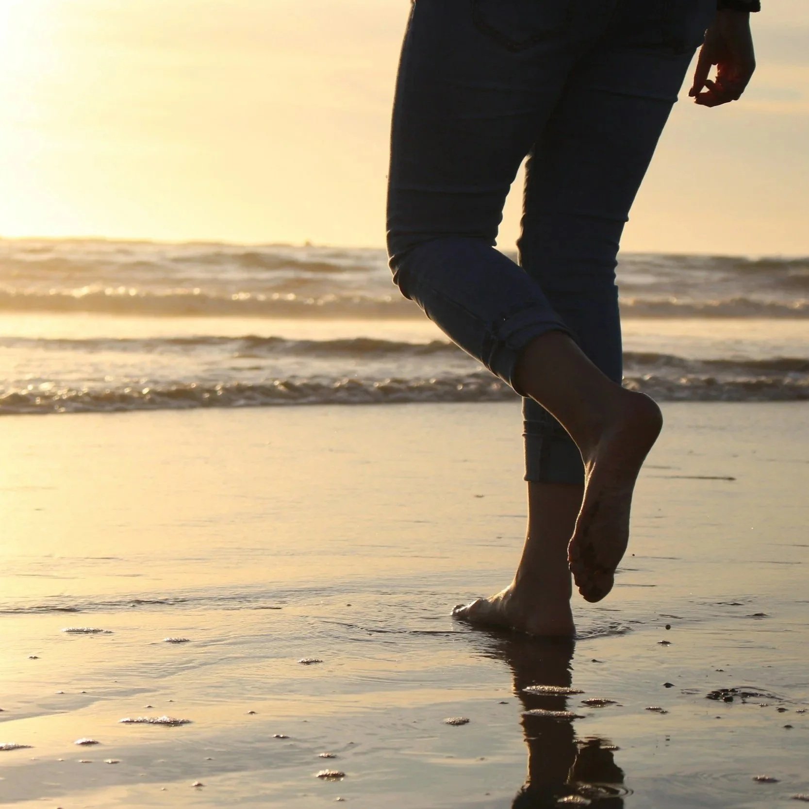 Person walking barefoot on the beach at sunset, with the ocean and waves in the background.