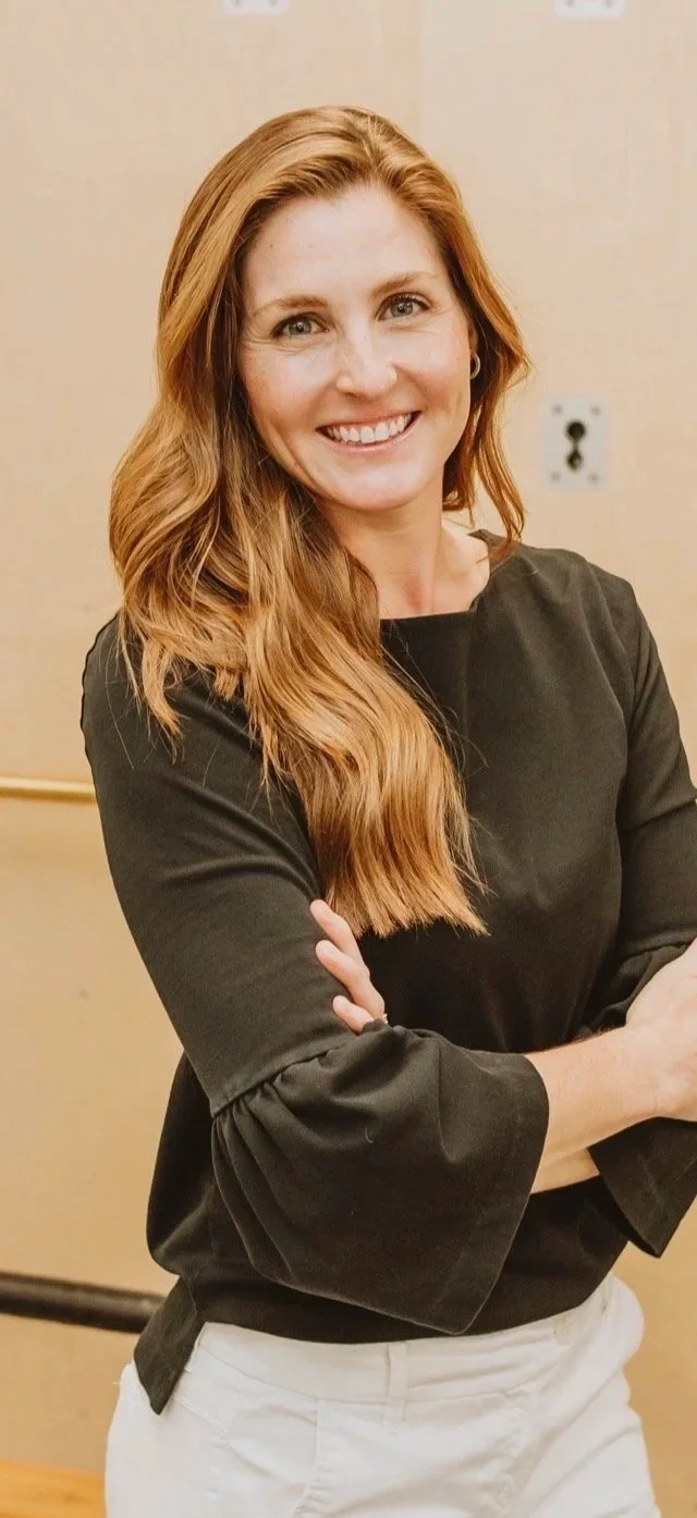 A woman with long, wavy red hair smiling and crossing her arms, wearing a black top and light-colored pants, standing in a room with beige walls.