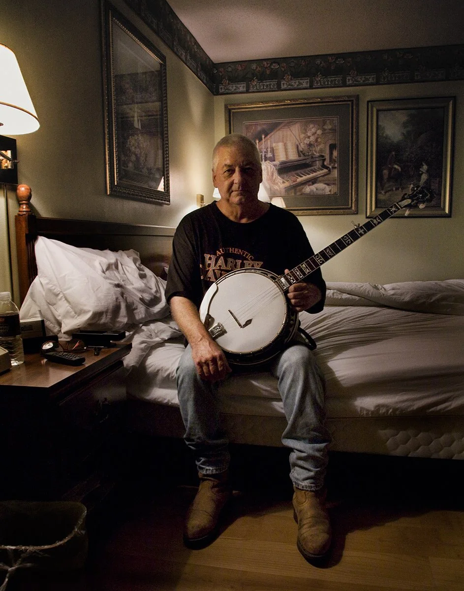 Man playing banjo in a motel room during a road trip, editorial portrait by Michael Rudin