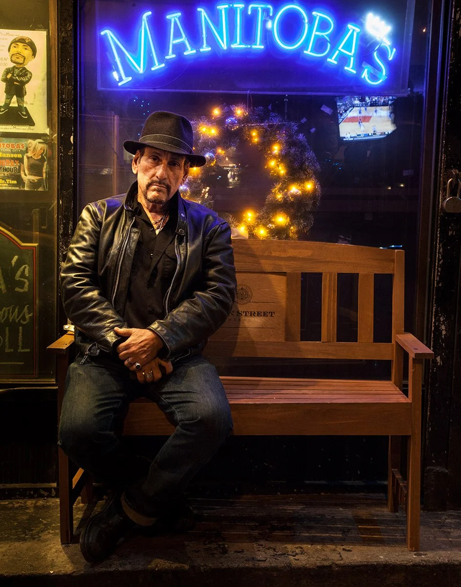 Dick Manitoba in a leather jacket and hat seated outside Manitoba's bar, editorial portrait by Michael Rudin