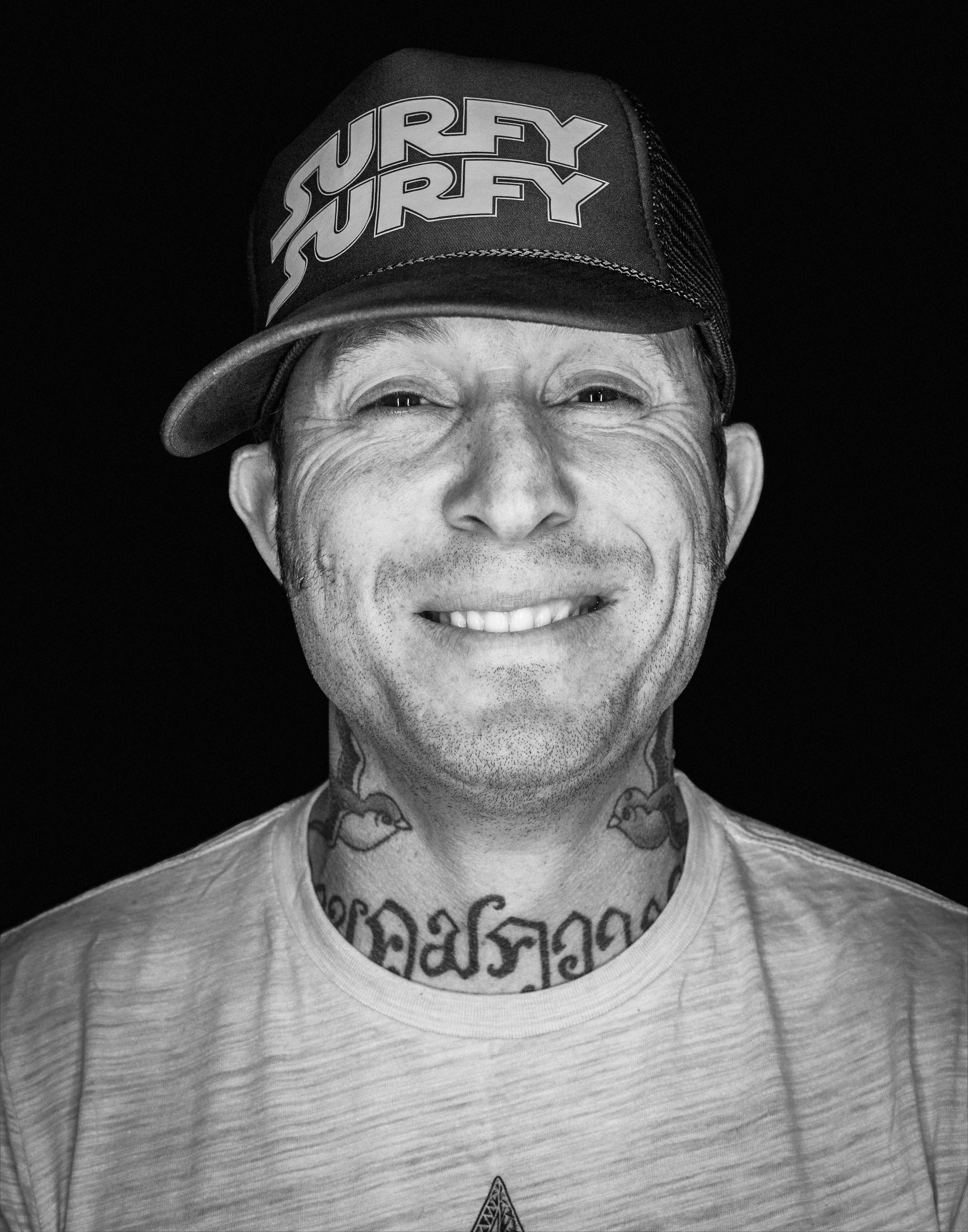 Black and white smiling portrait of a man in a trucker hat with neck tattoos, personal work by Michael Rudin
