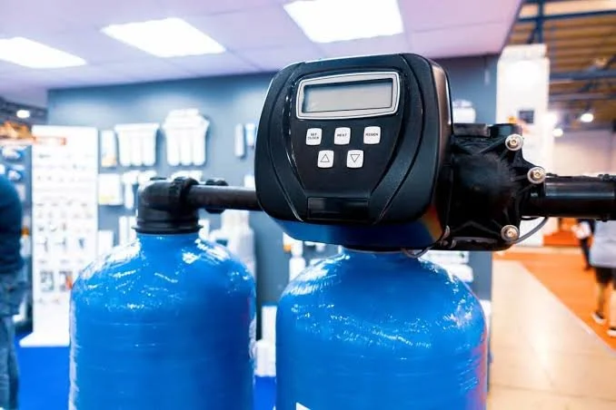 A digital water treatment control valve mounted on two large blue water bottles in an indoor setting.