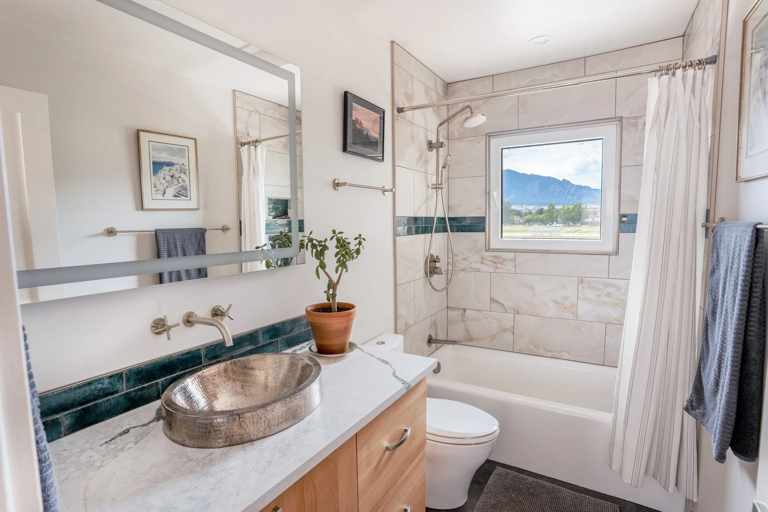 A small bathroom with a window showing a mountain view, a shower with a rainfall showerhead, a mirror, a plant in a pot, a copper sink, and towels hanging on the wall.