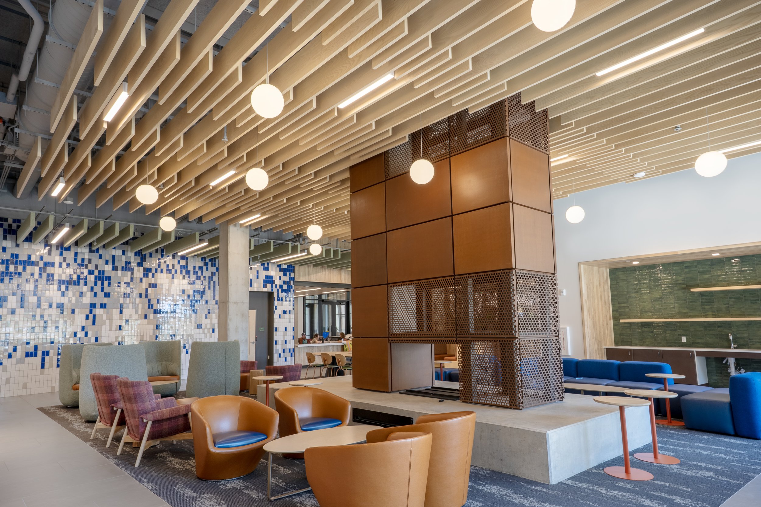 Modern interior space with a wooden slat ceiling, hanging spherical pendant lights, a tiled wall with blue and white mosaic pattern, brown and blue seating, and a reception area.