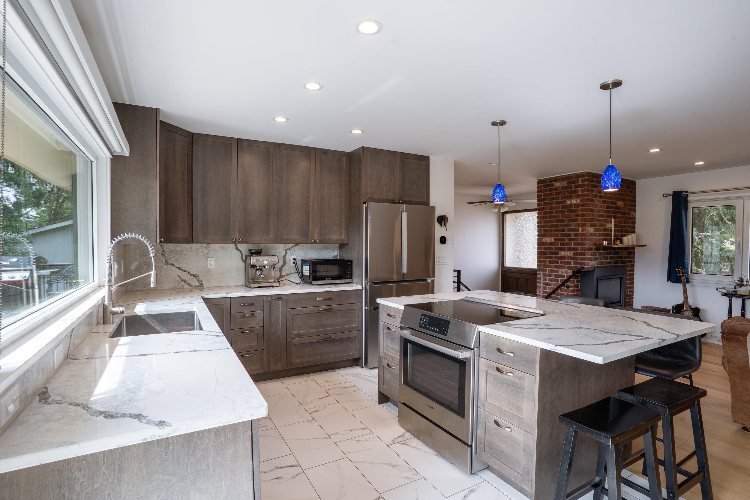 Modern kitchen with white and gray marble countertops, wood cabinets, stainless steel appliances, blue pendant lights, brick fireplace, and a ceiling fan.
