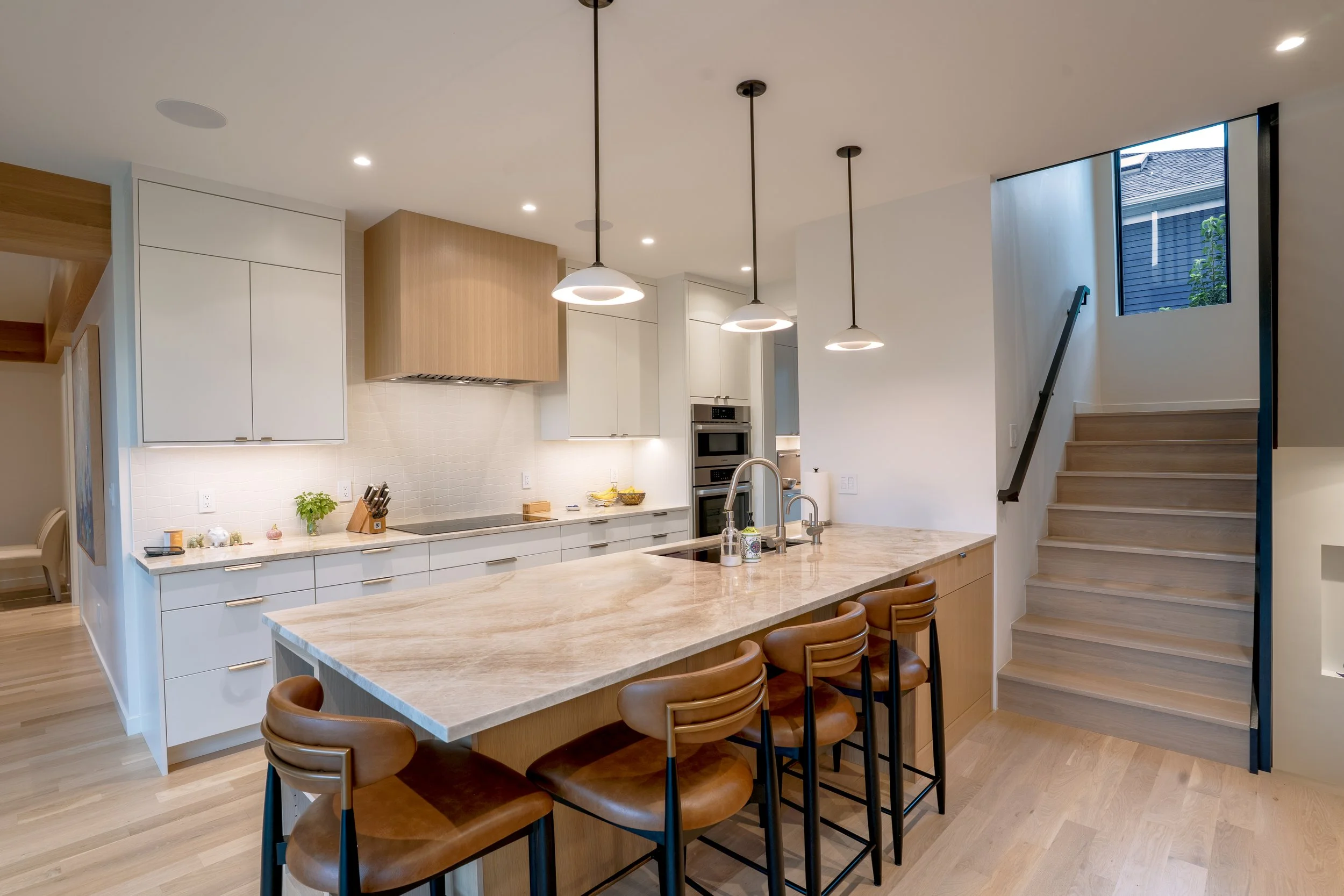 Modern kitchen with a light-colored marble island, brown bar stools, white cabinetry, a window near the staircase, pendant lights overhead, and stainless steel appliances.