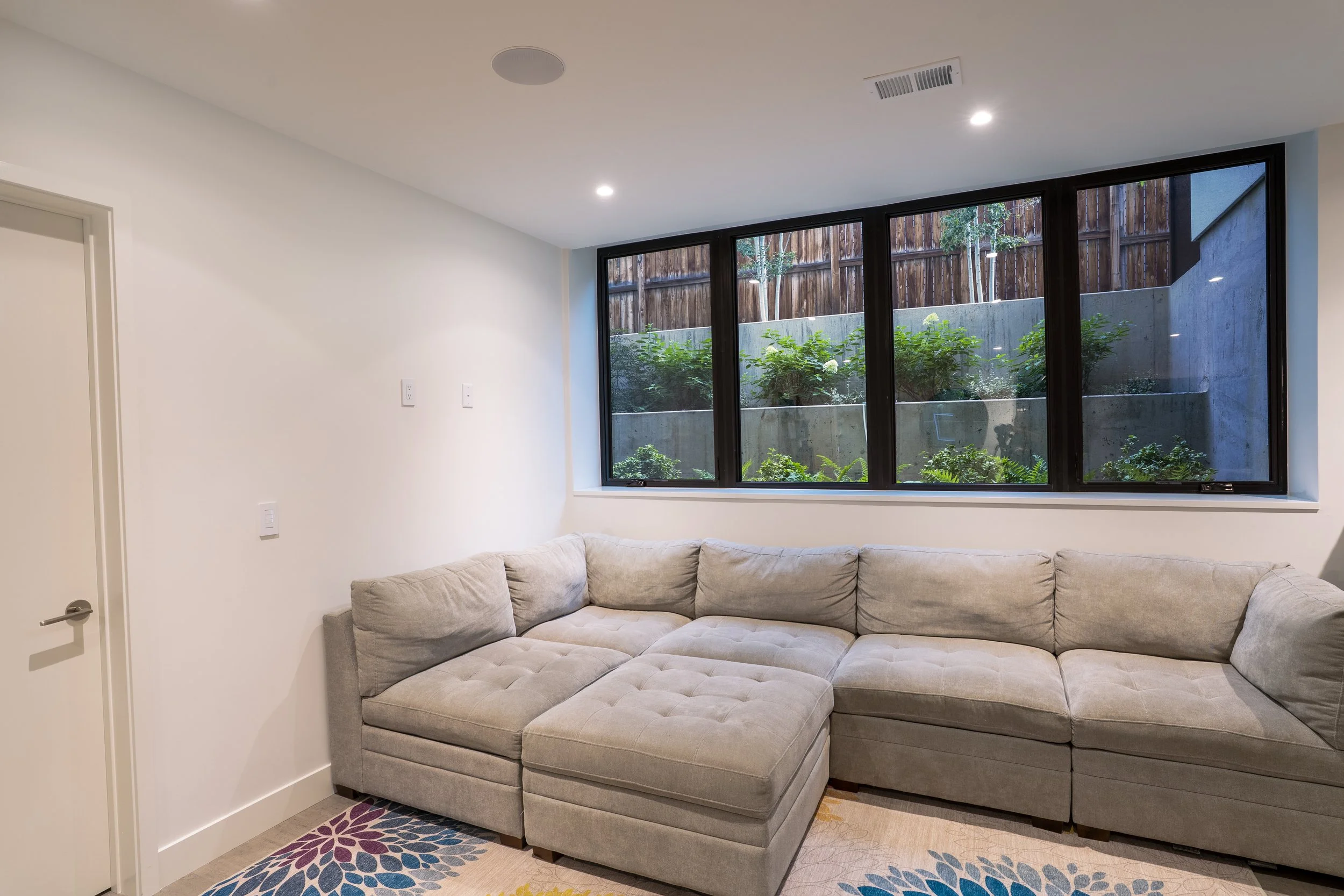 A cozy living room with a large gray sectional couch, a colorful floral rug, a big window looking out to a small patio with outdoor plants and a wooden fence, white walls, and recessed ceiling lights.
