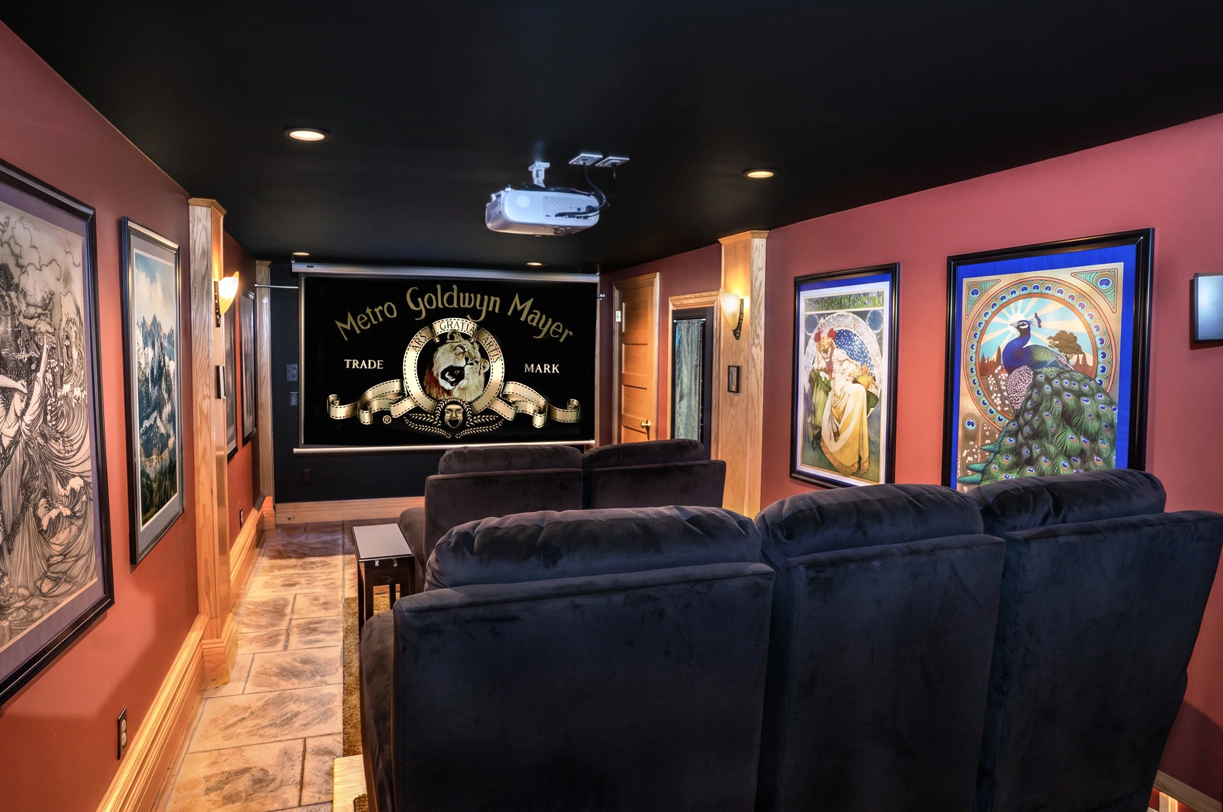 A home theater room with red walls, wooden trim, and artwork on the walls. There is a ceiling-mounted projector facing a black screen that displays the Metro Goldwyn Mayer logo. The seating consists of dark plush sofas arranged facing the screen.