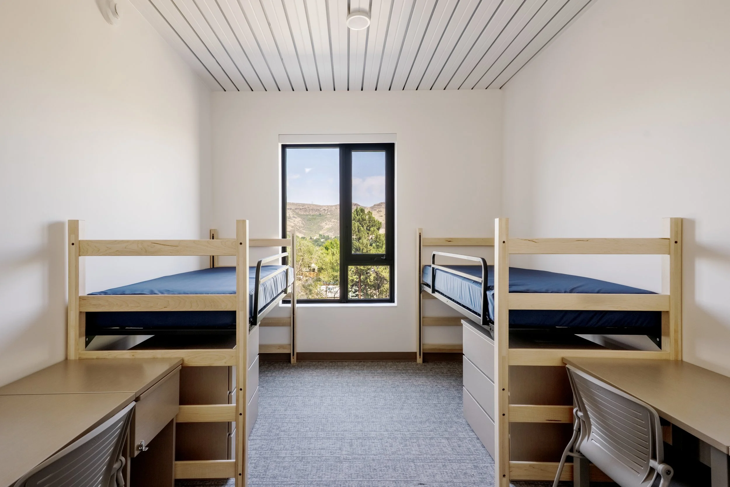 A dormitory room with two wooden bunk beds with blue mattresses, a desk and chair under the beds, a window with a landscape view of trees and hills, and a slatted ceiling.