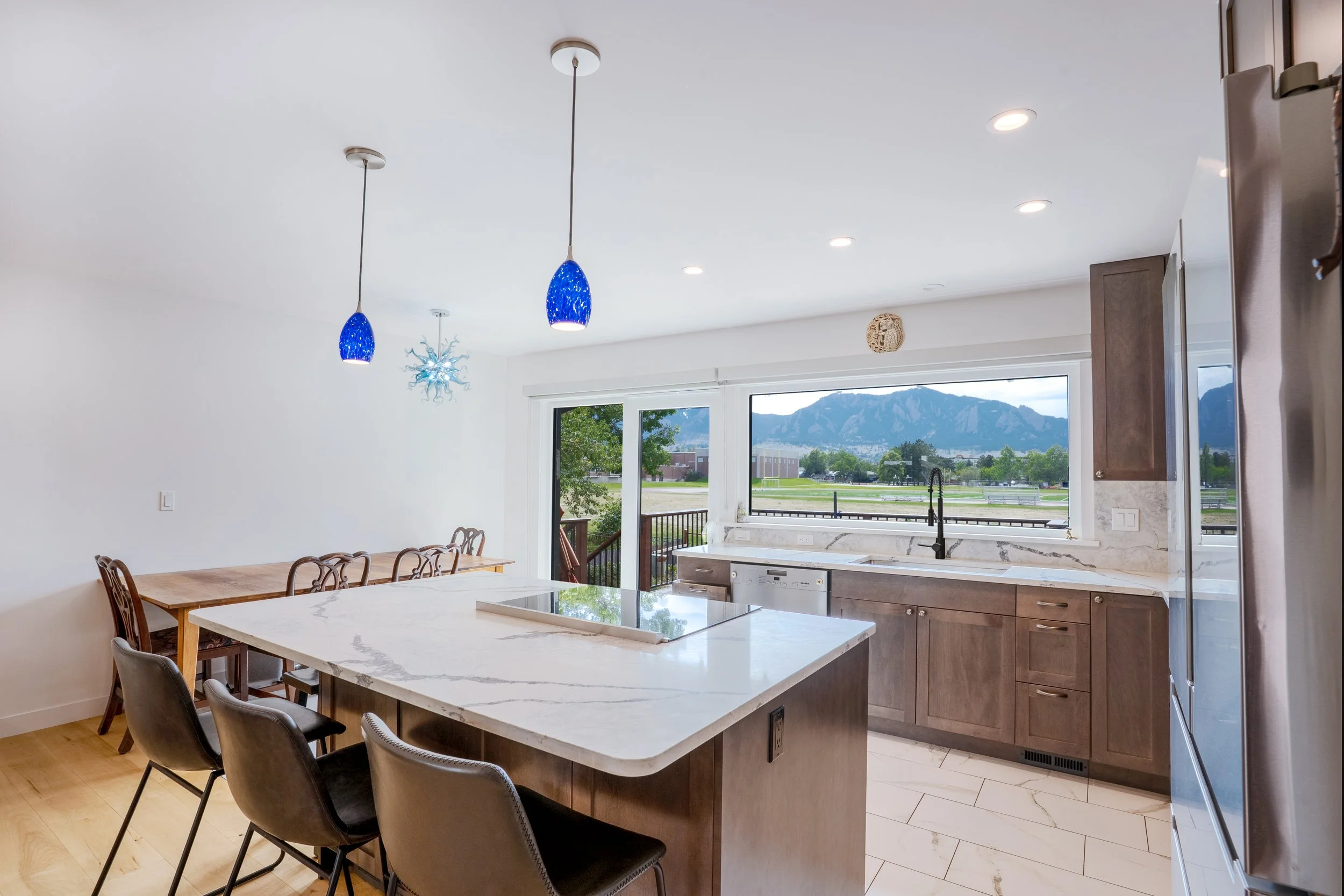 Modern kitchen with a large island, black chairs, wooden cabinets, marble countertops, and a window showing mountains outside. Hanging blue pendant lights and a snowflake-shaped chandelier are visible.