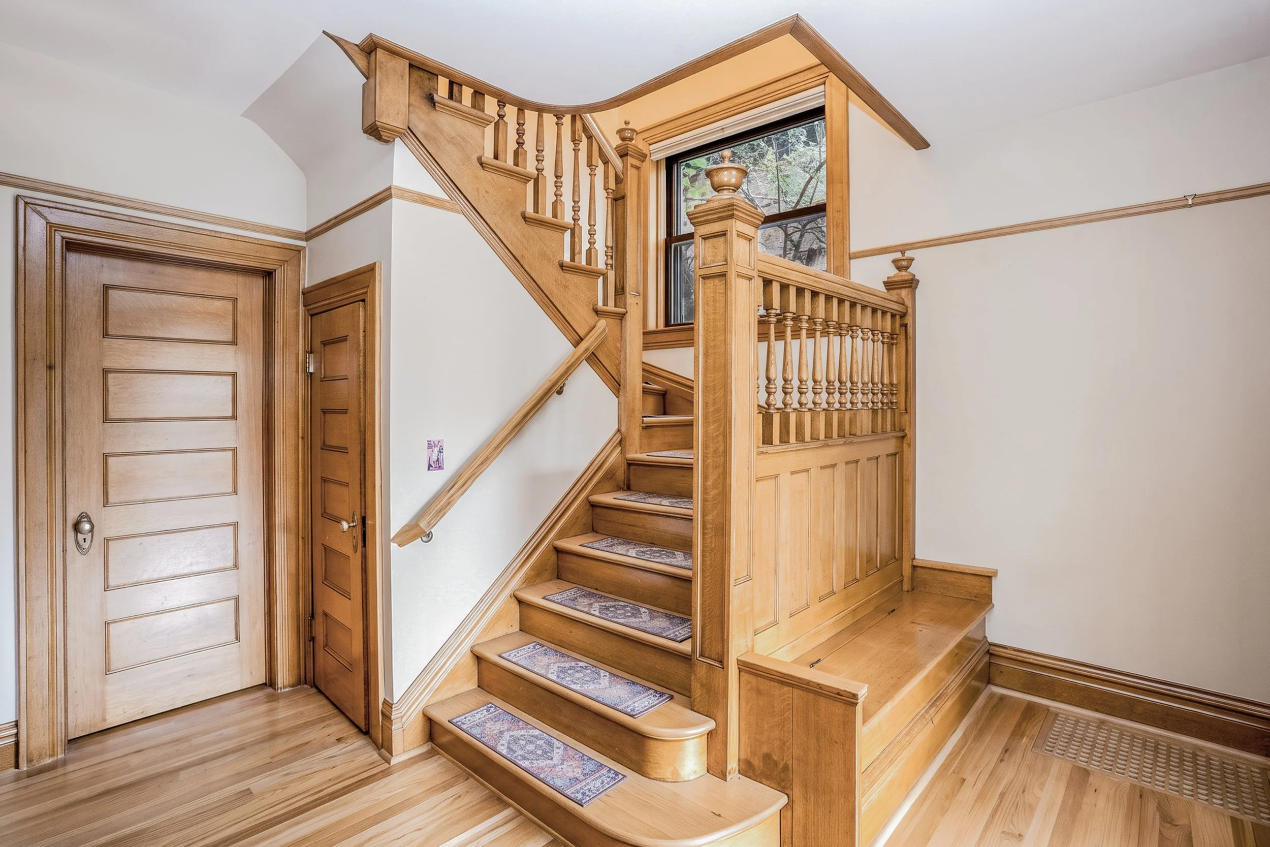 Wooden staircase with detailed spindles and handrail inside a house, with a large window and hardwood floors.