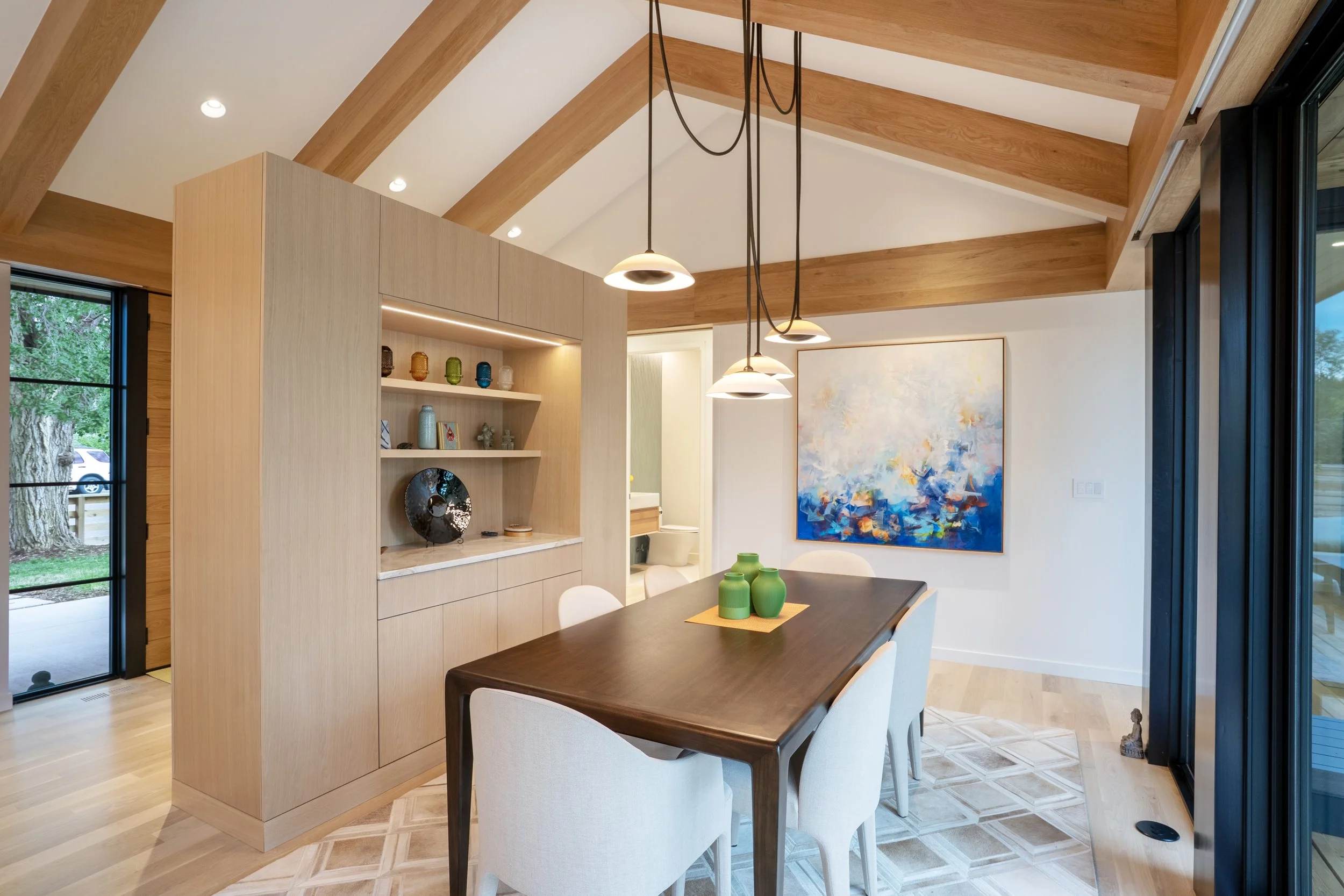 Interior view of a modern dining and living space with high wooden vaulted ceiling, pendant lighting, and colorful decorative items on shelves. Large window and sliding door reveal greenery outside.