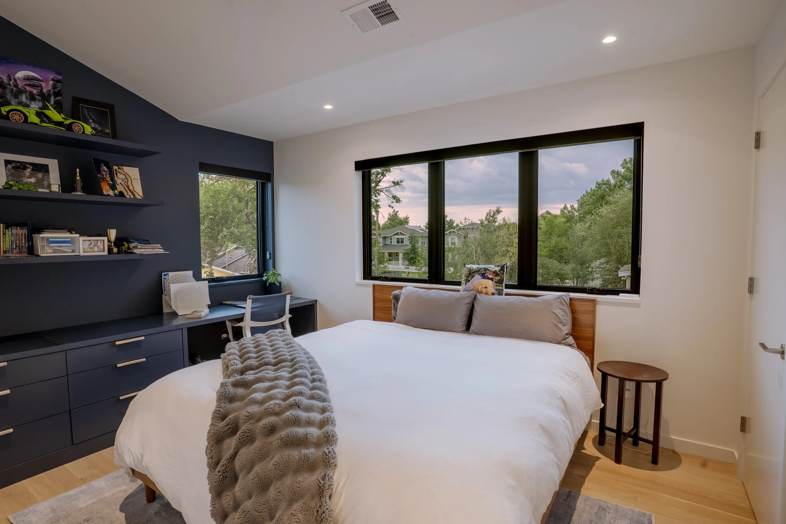 Modern bedroom with large windows showing a neighborhood view, a bed with gray pillows and a beige blanket, a navy blue dresser with shelves, artwork and books, a small side table, and a dog on the bed.