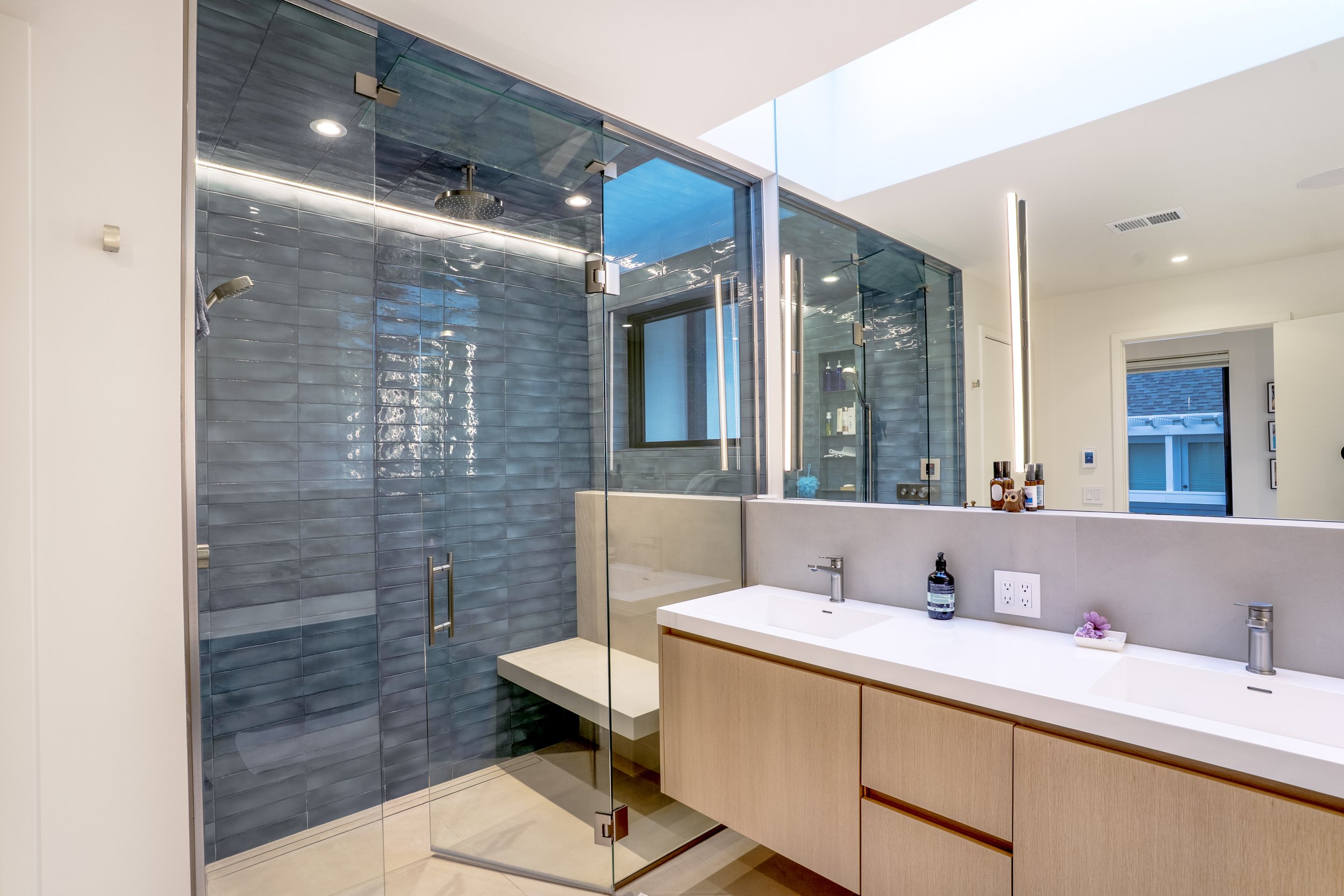Modern bathroom with a glass-enclosed shower with blue tiles, a white vanity with a sink, and a window with a view of neighboring houses.